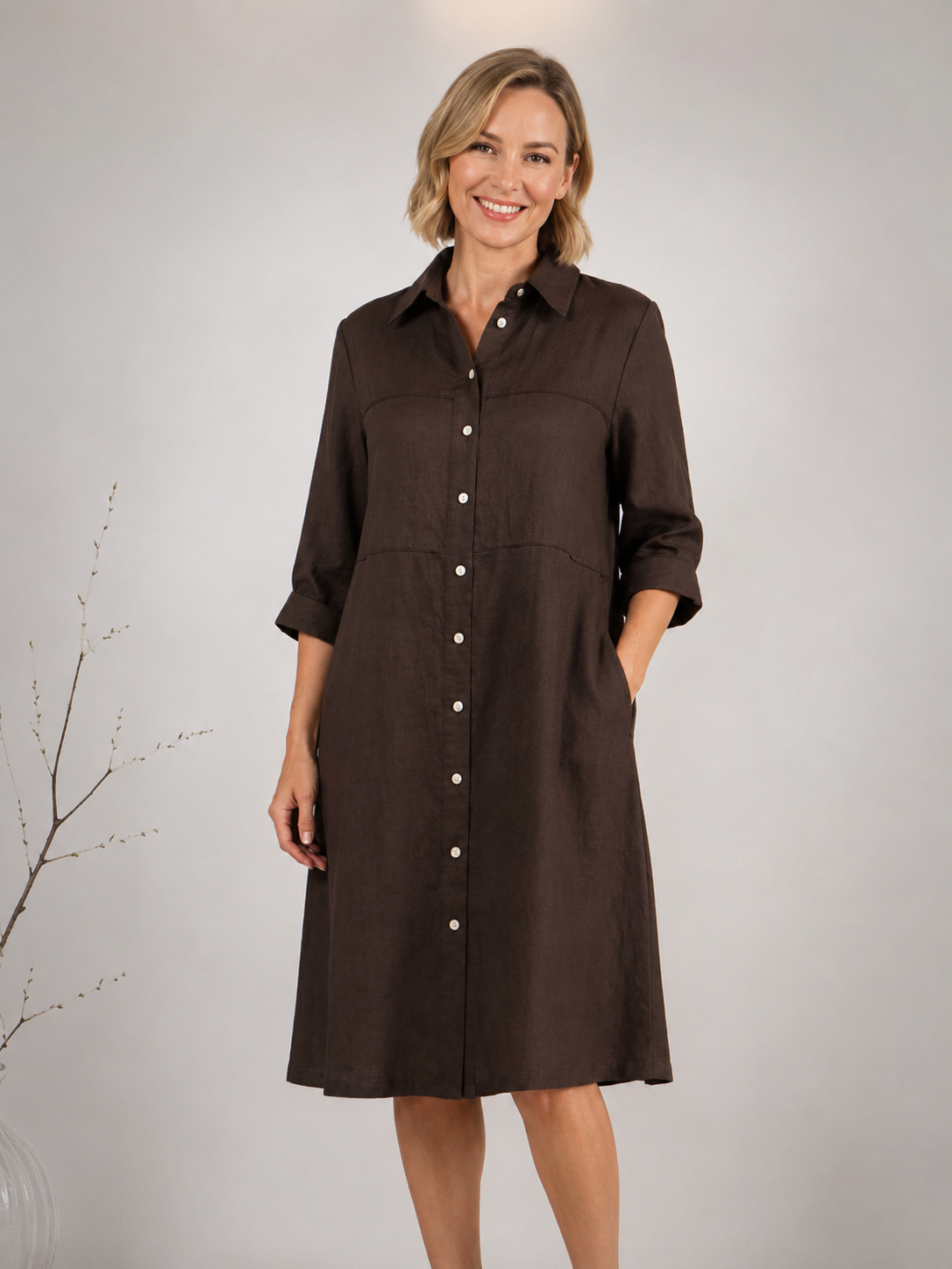 A woman modeling an espresso button-down See Saw shirtmaker dress with three-quarter sleeves and side pockets, standing against a neutral light gray studio background.