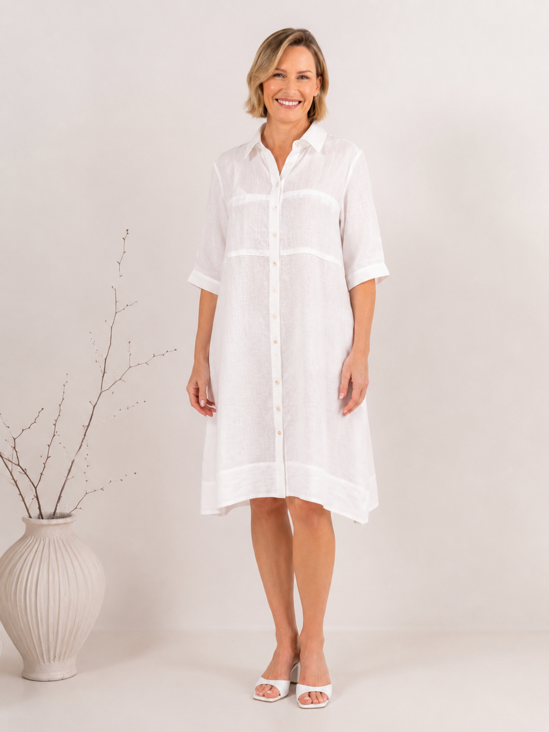 A woman modeling an white linen button-down See Saw shirtmaker dress with three-quarter sleeves and side pockets, standing against a neutral light gray studio background.