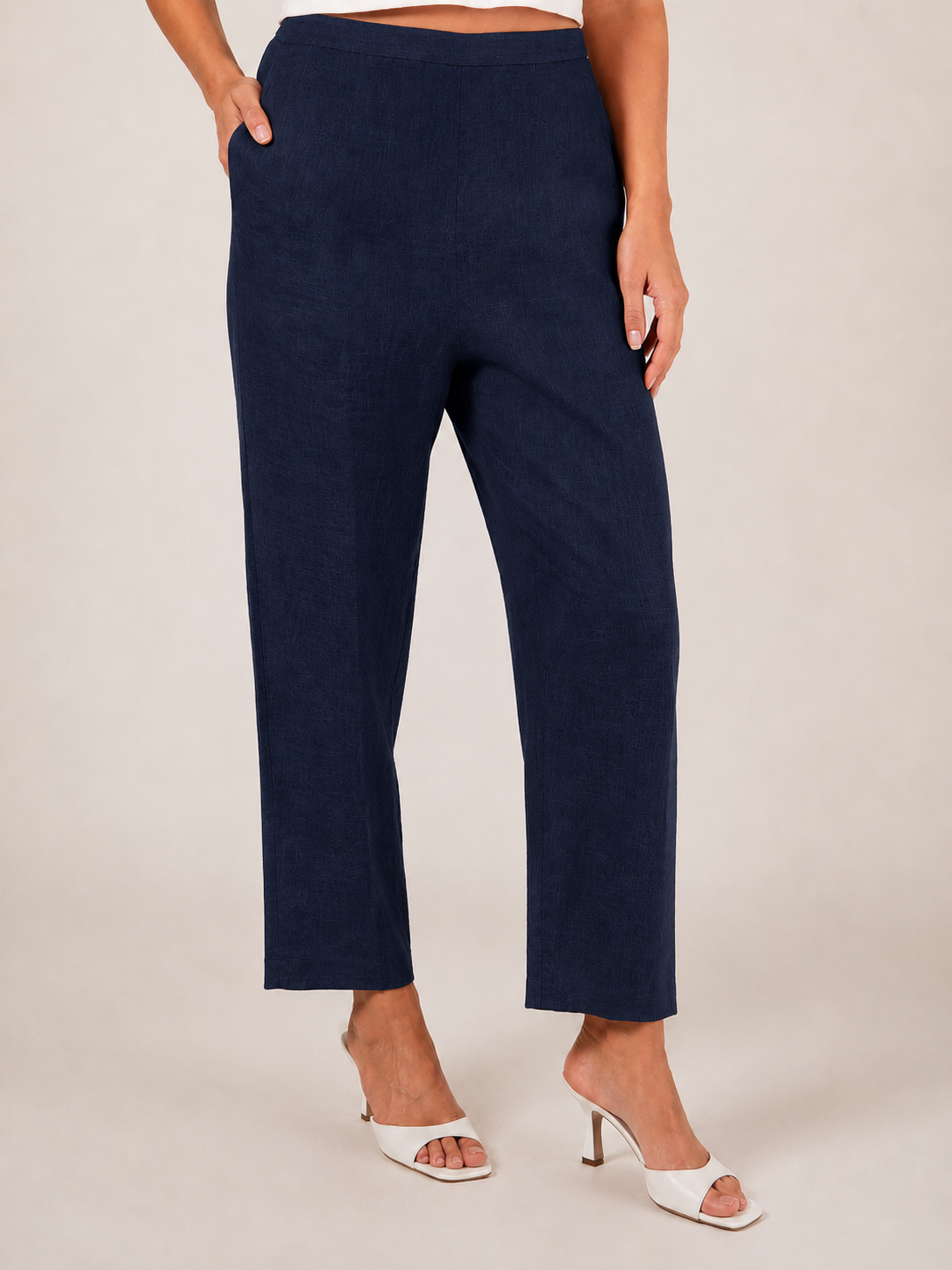 A person wearing navy linen  See Saw 7/8 pants with a relaxed straight-leg fit, paired with white heeled slide sandals, photographed against a plain light beige studio background.