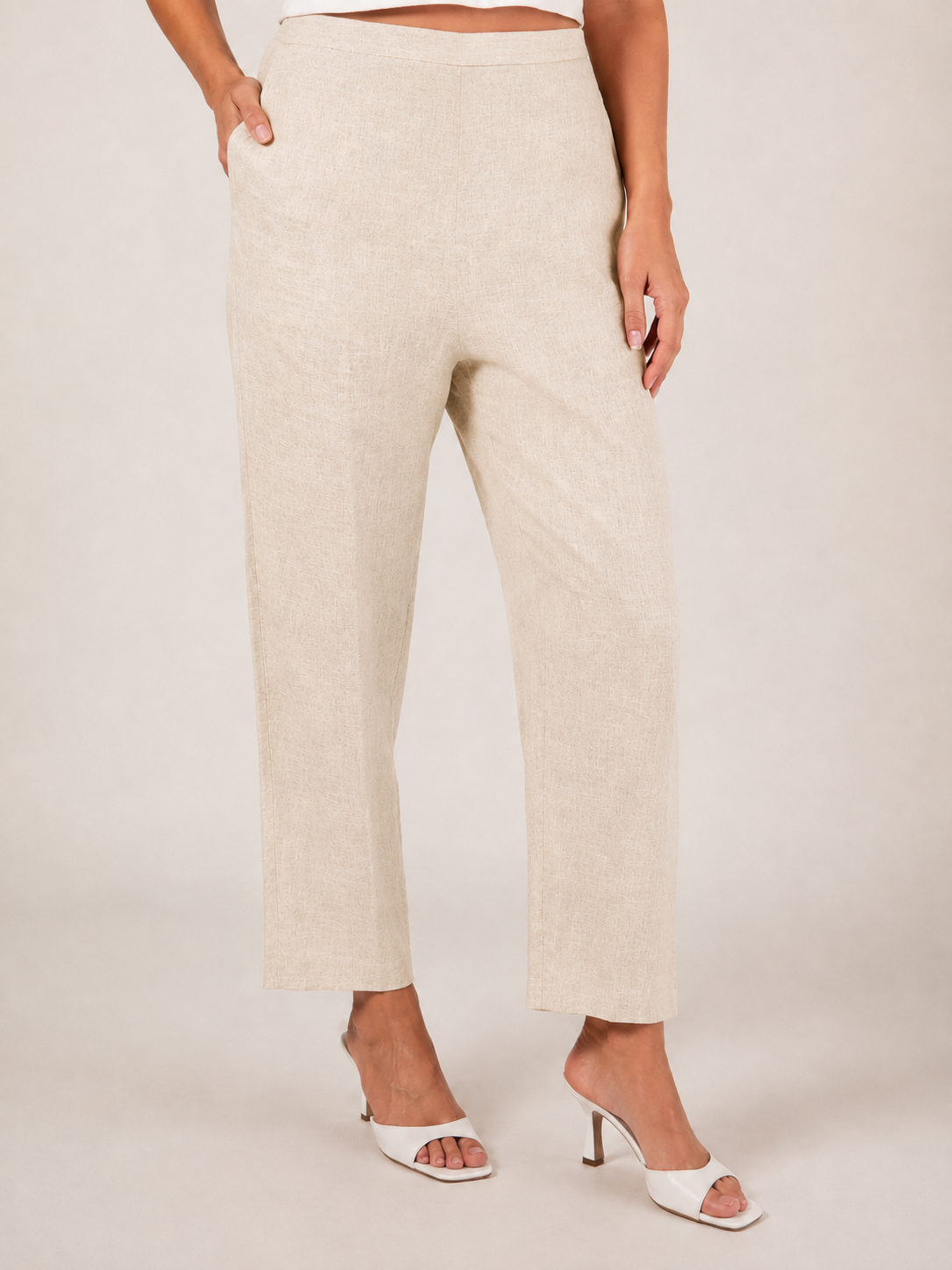 A person wearing natural linen  See Saw 7/8 pants with a relaxed straight-leg fit, paired with white heeled slide sandals, photographed against a plain light beige studio background.