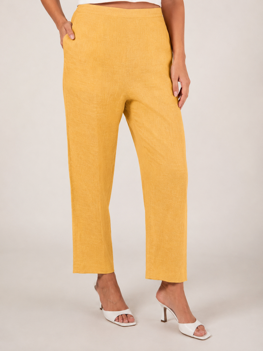 A person wearing marigold linen  See Saw 7/8 pants with a relaxed straight-leg fit, paired with white heeled slide sandals, photographed against a plain light beige studio background.