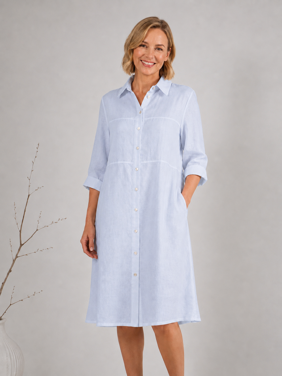 A woman modeling a blue linen button-down See Saw shirtmaker dress with three-quarter sleeves and side pockets, standing against a neutral light gray studio background.