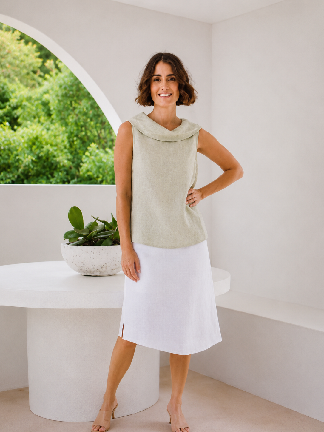A woman modeling a two-piece See Saw linen set consisting of a light sage green sleeveless cowl-neck top and a white midi skirt, standing in a bright, modern white room with an arched window view of green foliage.