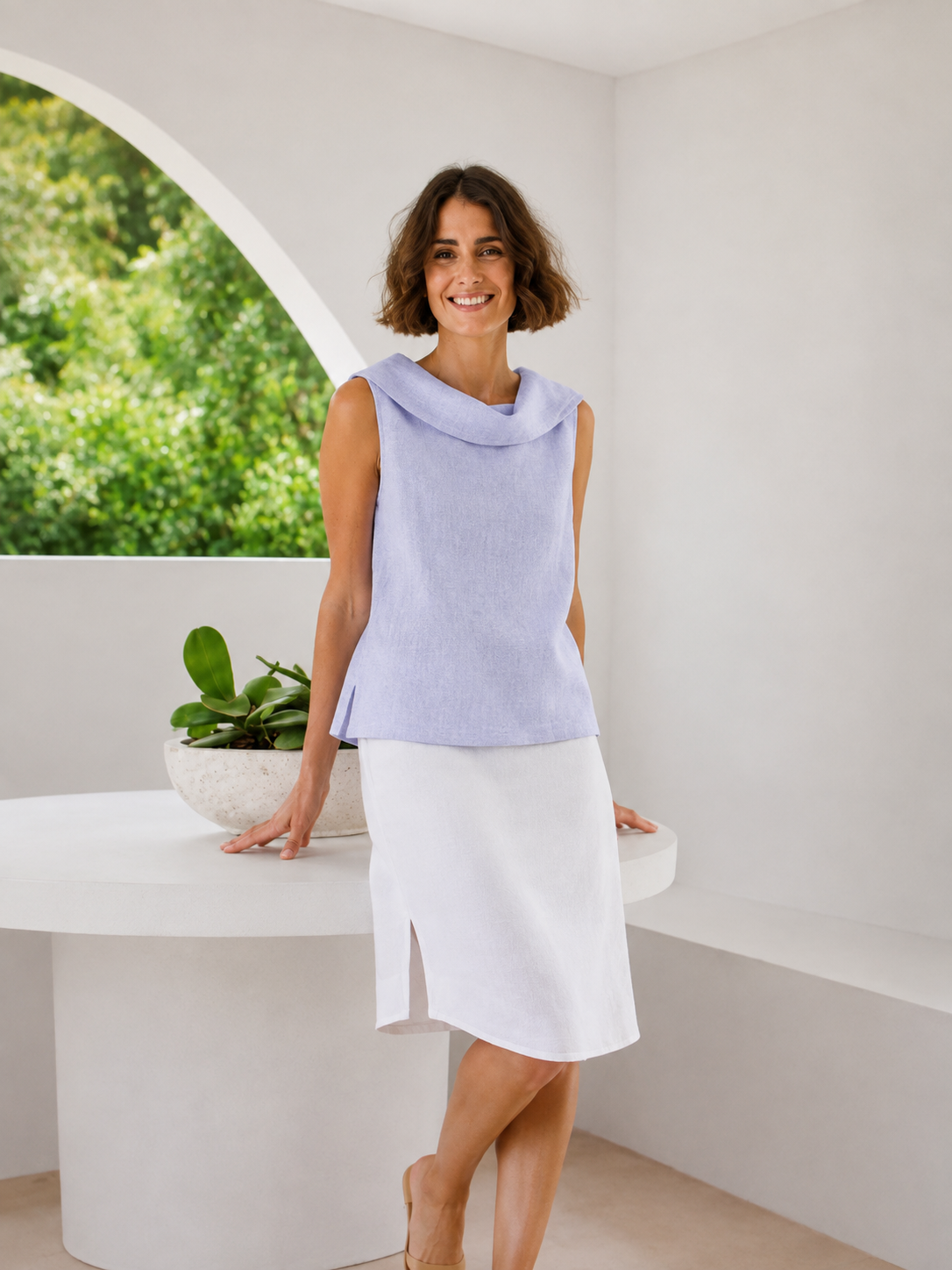 A woman modeling a sleeveless light sky blue See Saw linen cowl-neck top and a white skirt, standing in a bright, minimalist white room with a curved white table and a potted plant.