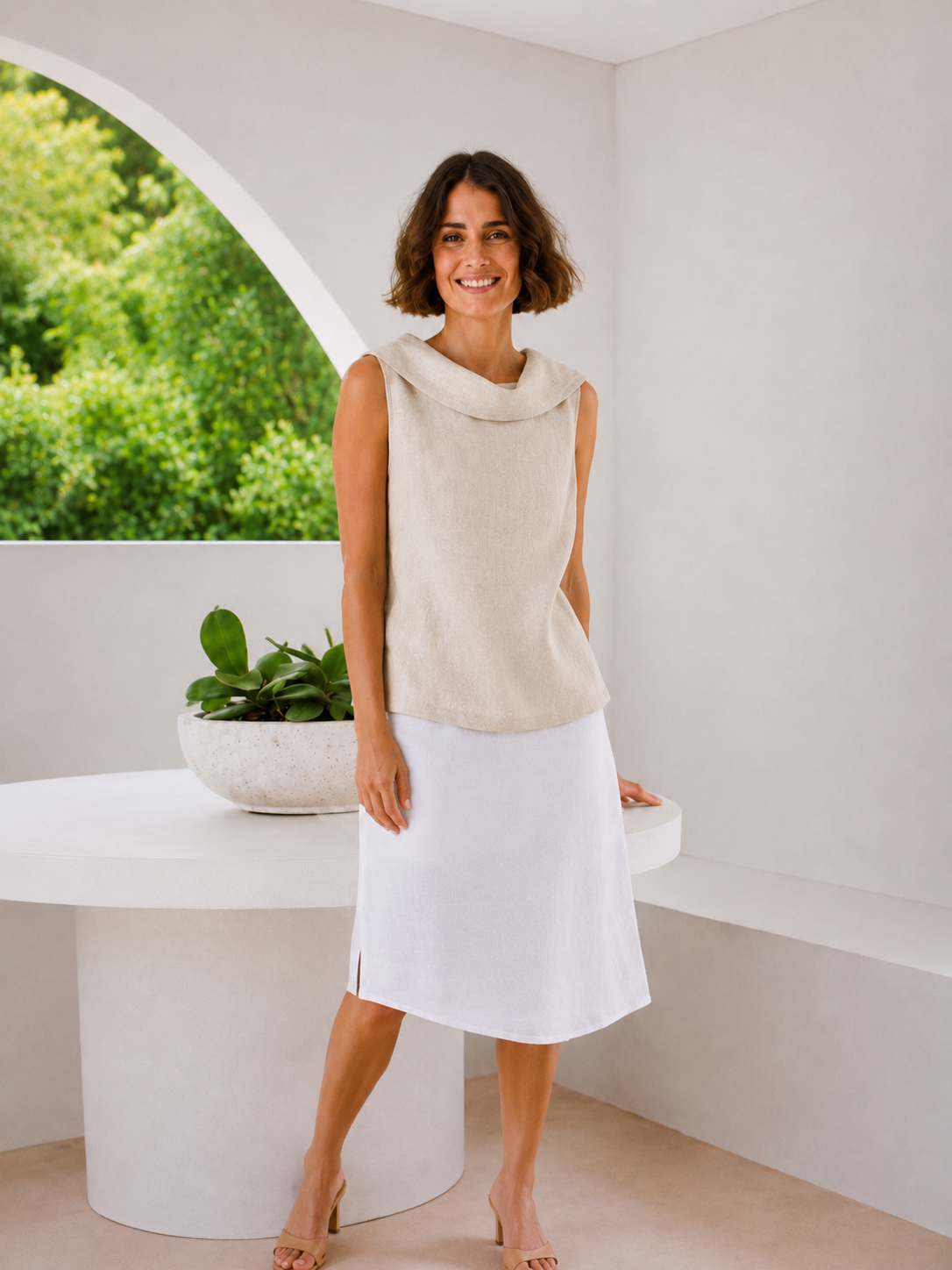 A woman modeling a sleeveless cowl-neck top in a light beige linen-like fabric, paired with a white midi skirt, standing in a bright, modern white room with a large arched window showing green foliage outside.