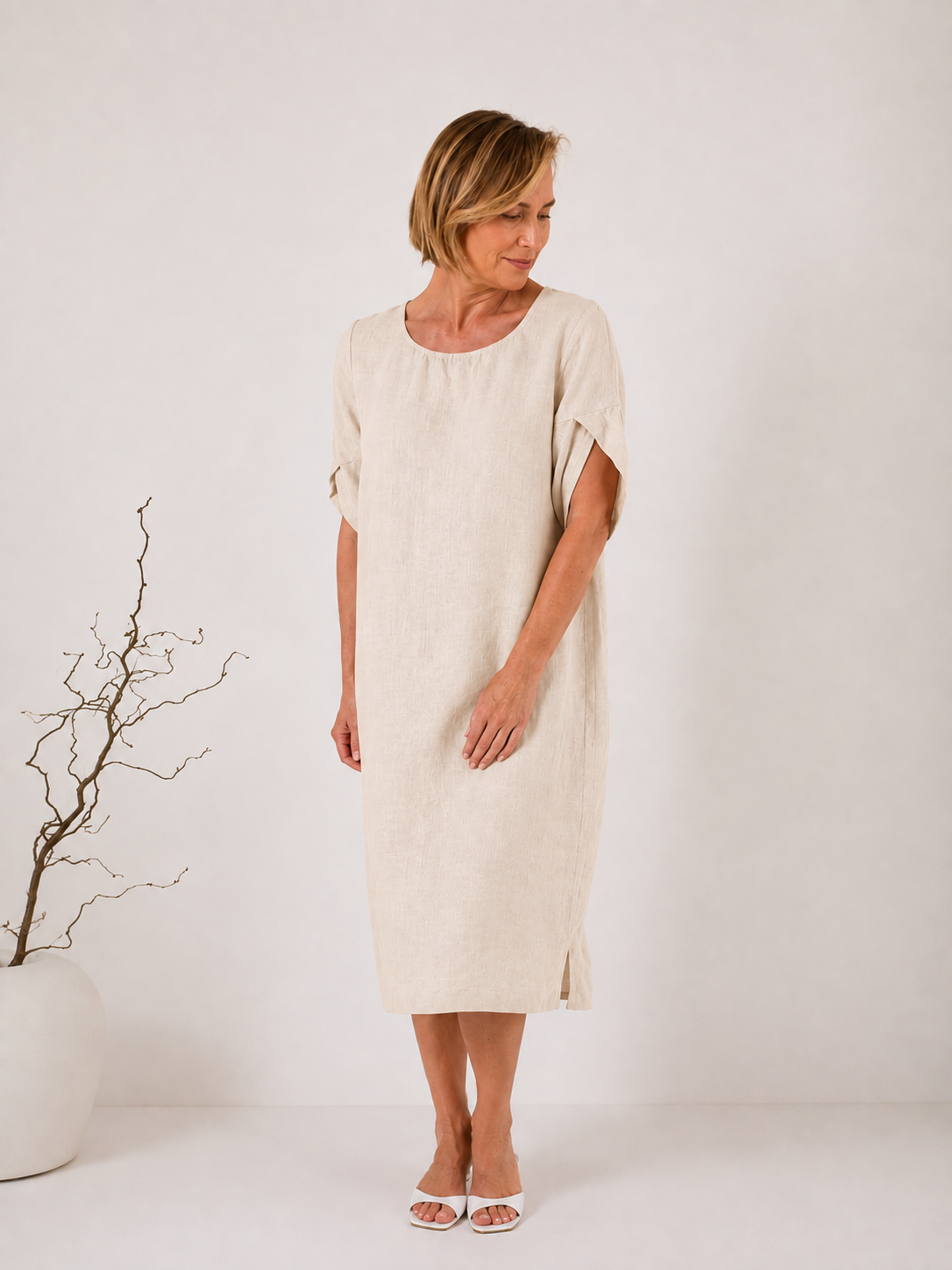 A woman modeling a short-sleeved, knee-length linen dress in a light cream or off-white color, standing against a plain white studio background.