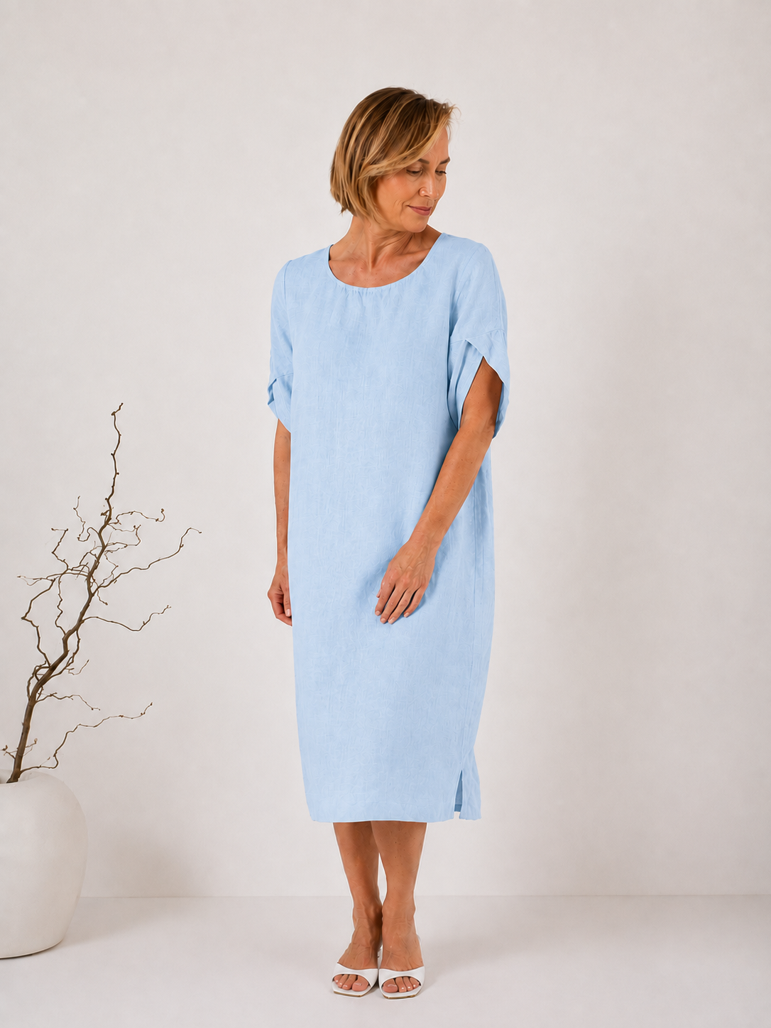 A woman wearing a light blue linen midi dress with short sleeves and a relaxed fit, standing in a minimalist studio setting with a white wall and a decorative vase of branches on the left.