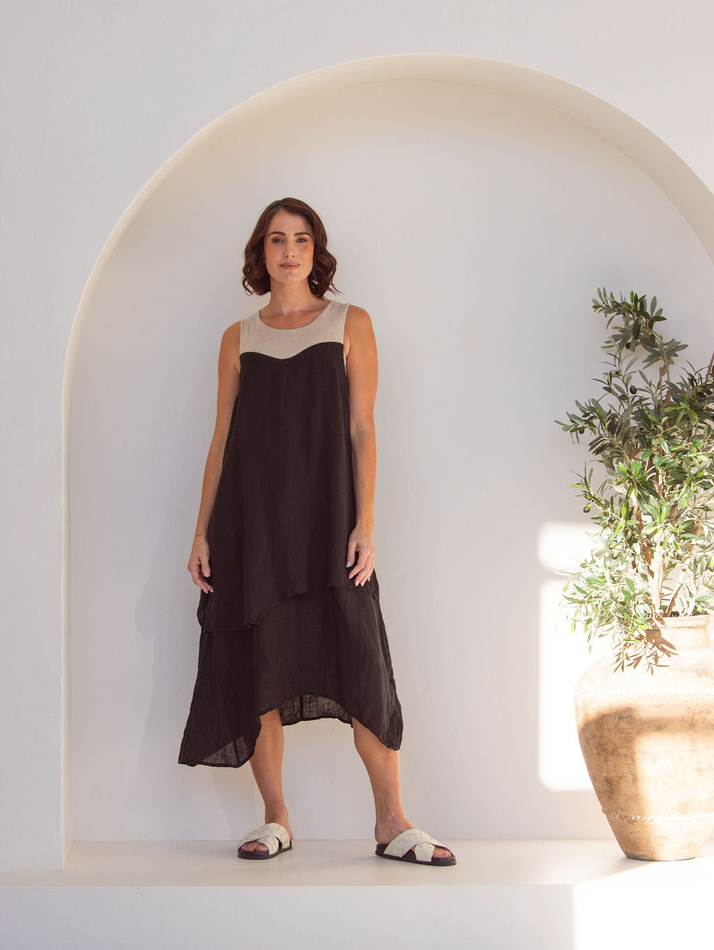 Woman wearing a black and beige linen See Saw midi dress. Standing next to a plant in an arch minimal background.