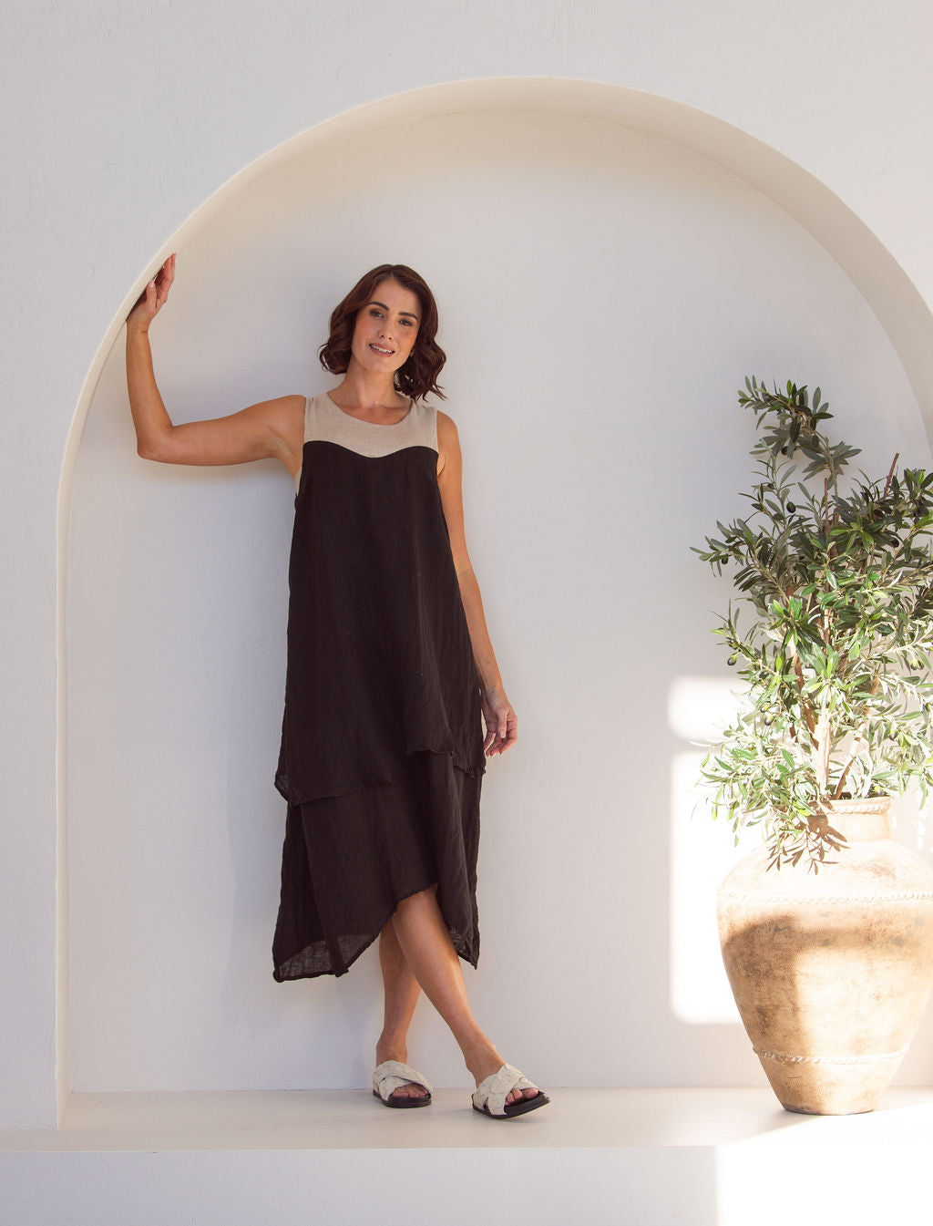 Woman wearing a black and beige linen See Saw midi dress. Standing next to a plant in an arch minimal background.