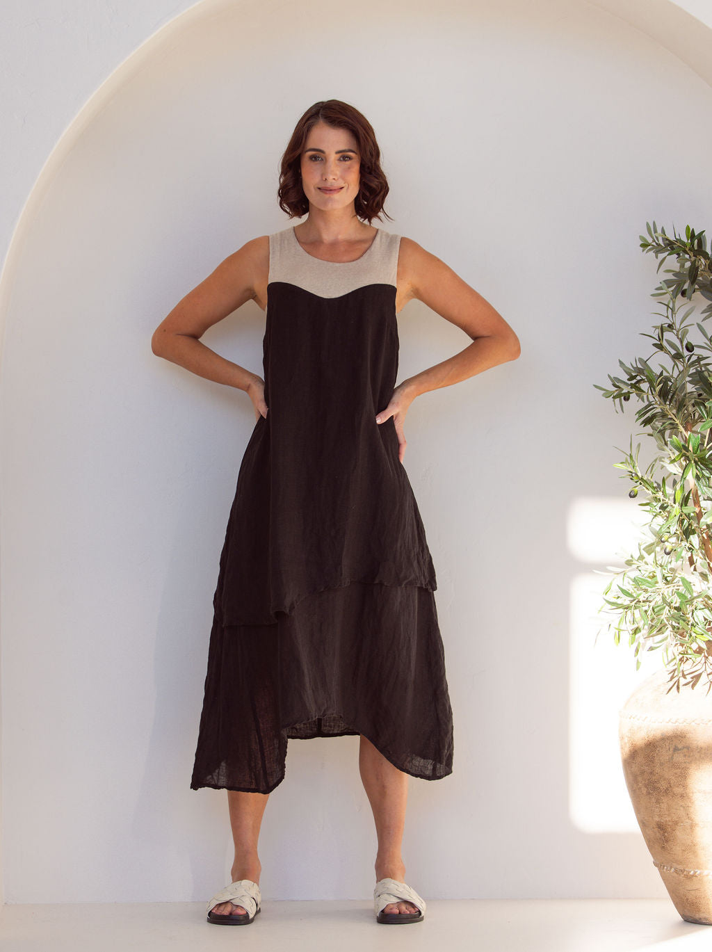 Woman wearing a black and beige linen See Saw midi dress. Standing next to a plant in an arch minimal background.