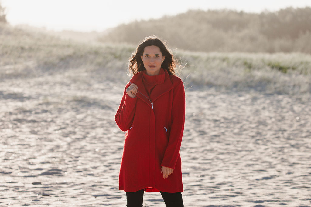 Woman wearing a boiled wool See Saw red coat on a beach with ocean and sky in the background