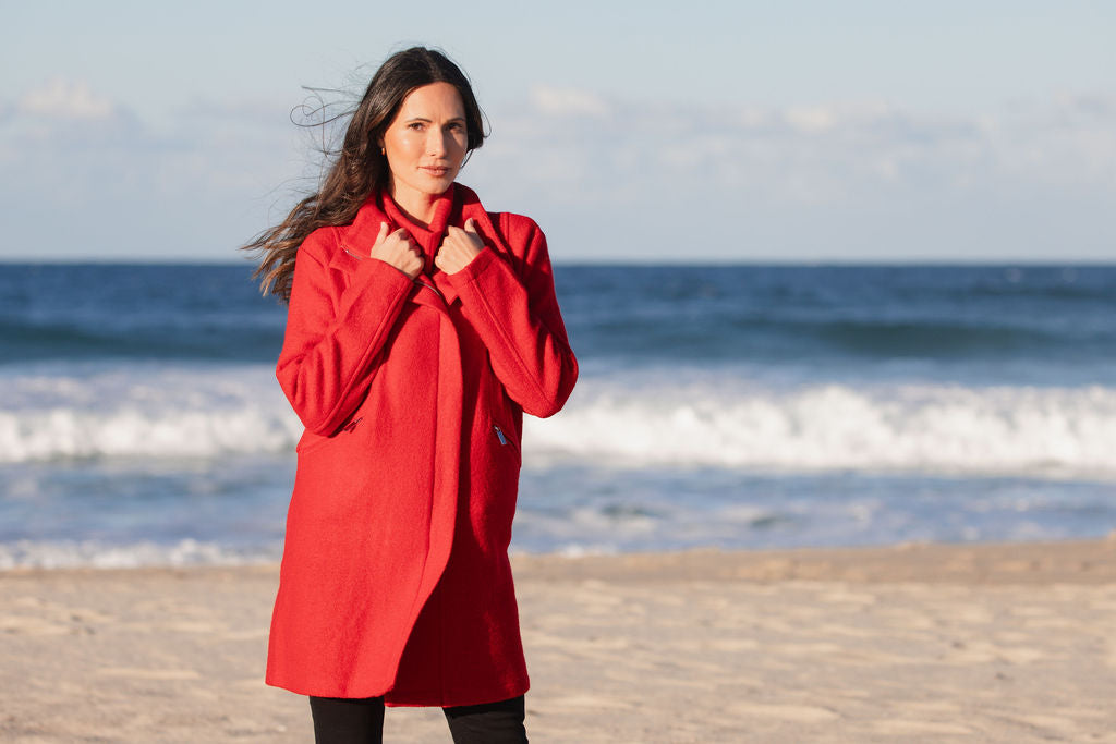 Woman wearing a boiled wool See Saw red coat on a beach with ocean and sky in the background