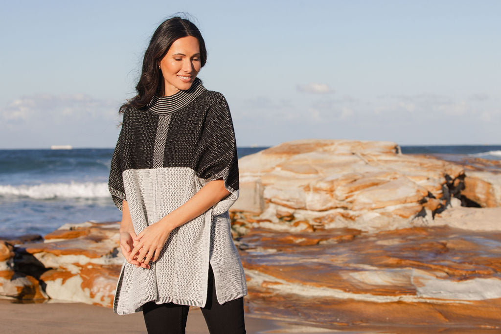 Woman wearing a See Saw wool  blend black and grey patterned poncho on a beach with ocean and rocks in the background