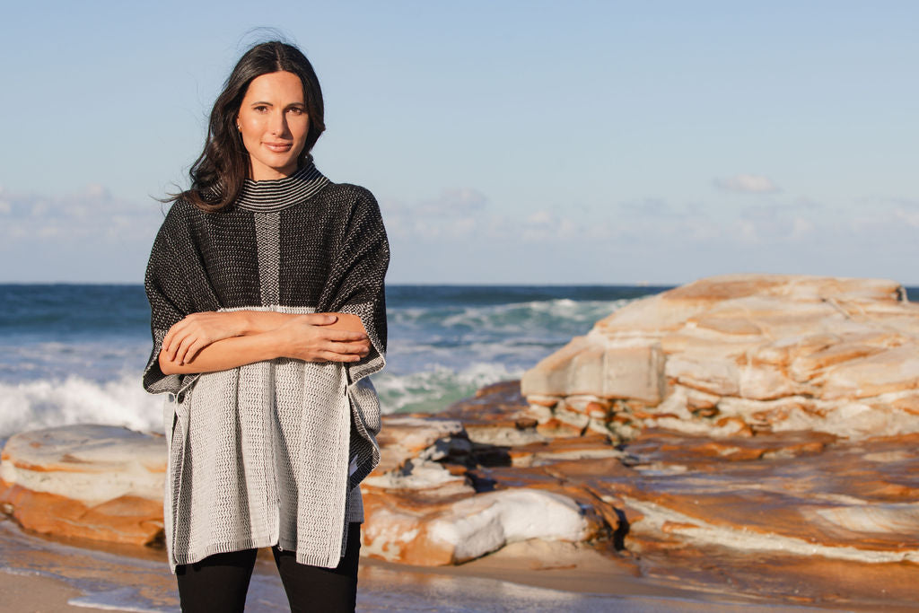 Woman wearing a See Saw wool  blend black and grey patterned poncho on a beach with ocean and rocks in the background