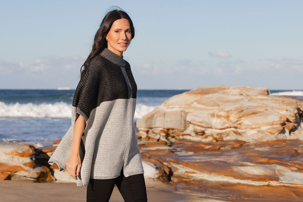 Woman wearing a See Saw wool  blend black and grey patterned poncho on a beach with ocean and rocks in the background