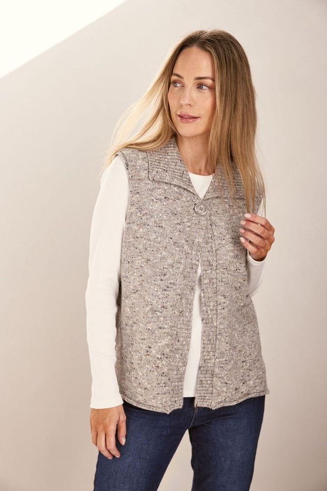 Woman wearing a patterned speckle Se Saw vest with a button over a white shirt and jeans on a plain background