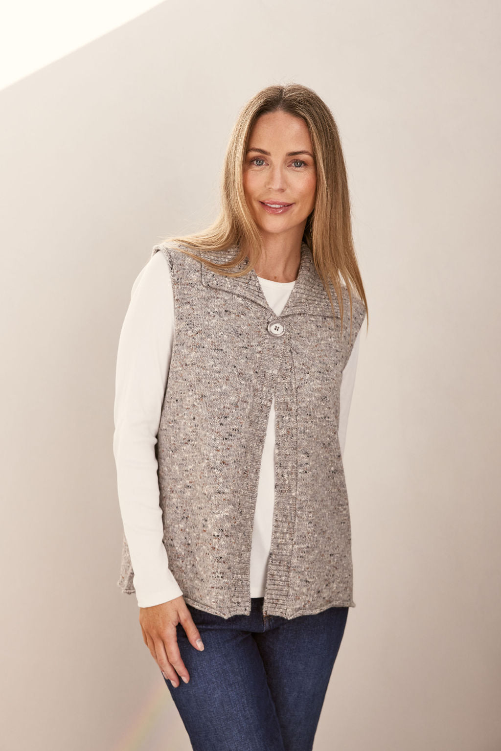 Woman wearing a patterned speckle Se Saw vest with a button over a white shirt and jeans on a plain background