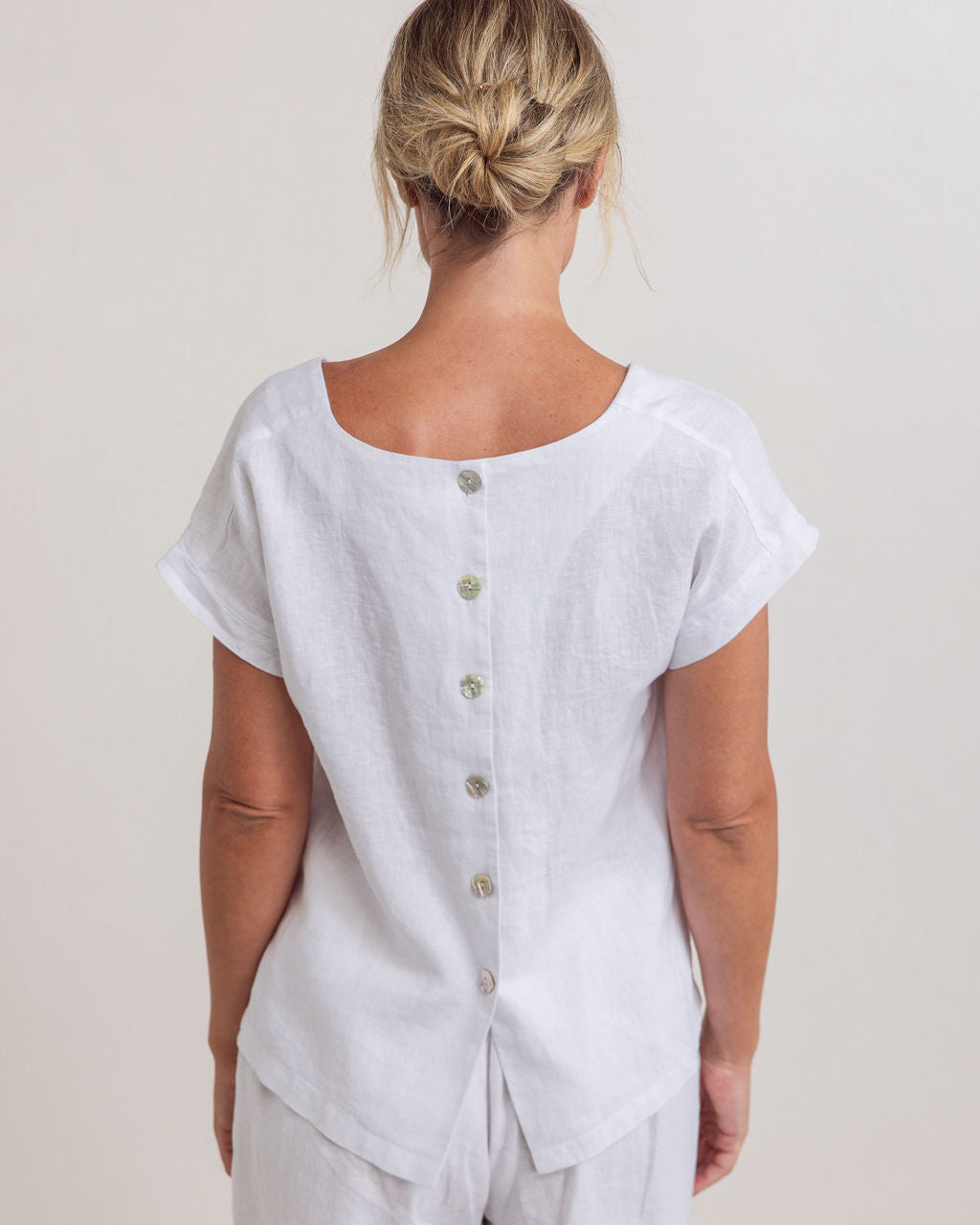 Woman in a white Linen See Saw top and pattern pants standing next to a potted plant in a minimalistic setting.