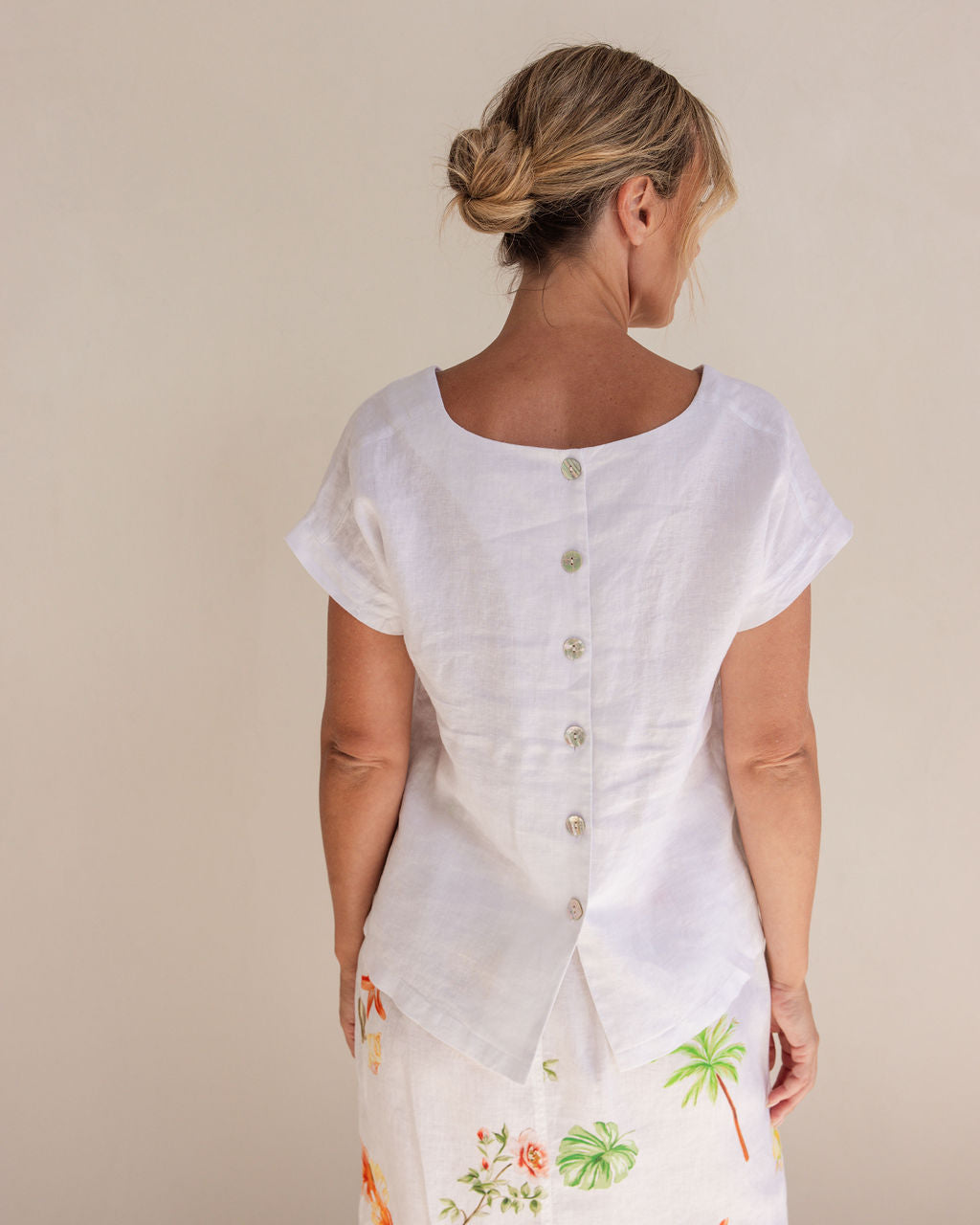 Woman in a white Linen See Saw top and white pants standing next to a potted plant in a minimalistic setting.