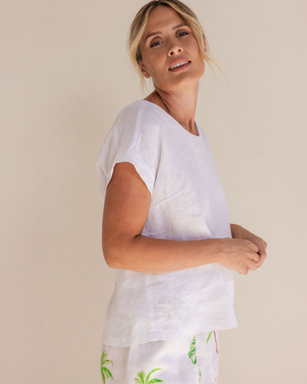 Woman in a white Linen See Saw top and white pants standing next to a potted plant in a minimalistic setting.