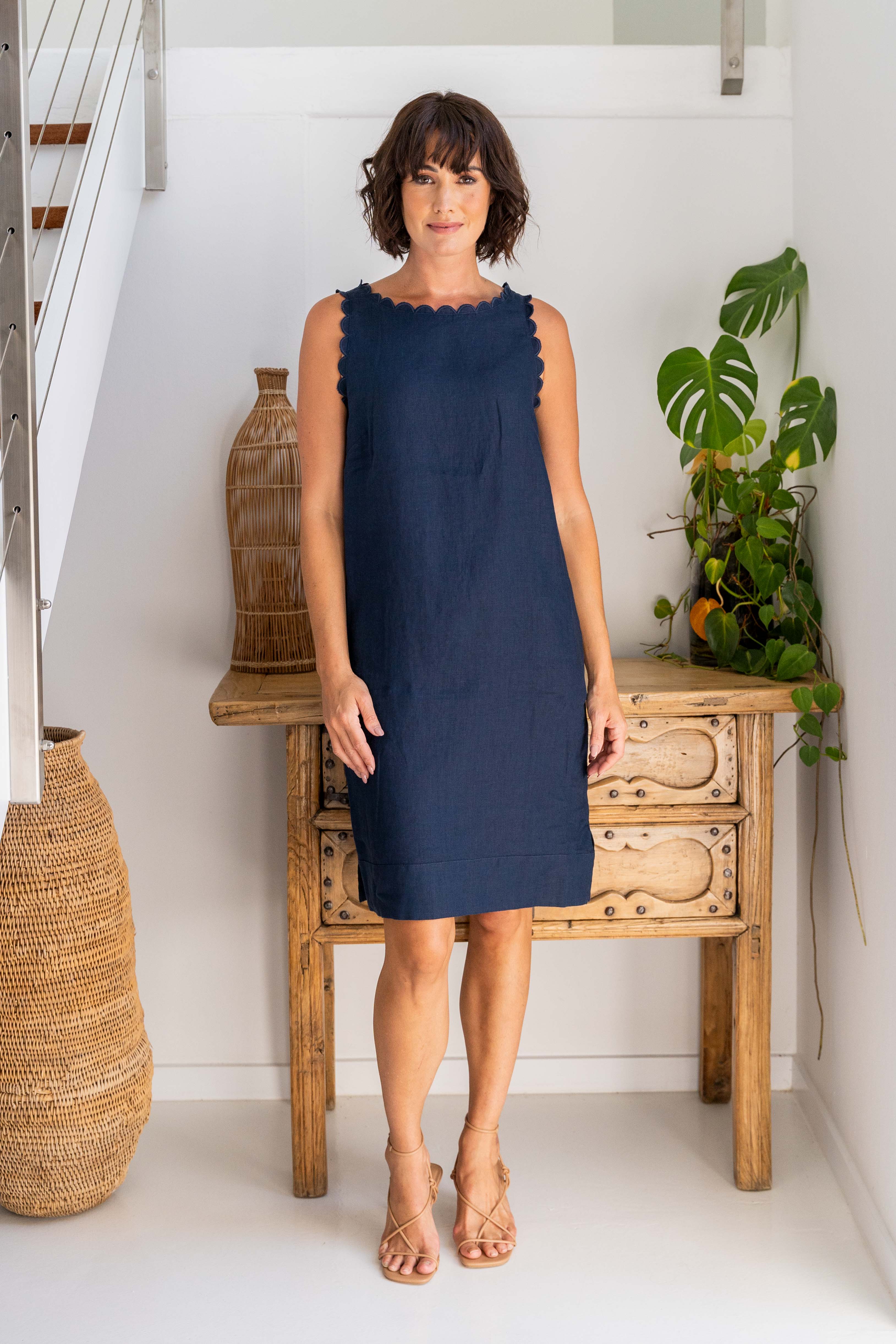 Woman wearing a navy linen See Saw dress against a white background with wooden drawers and a plant.