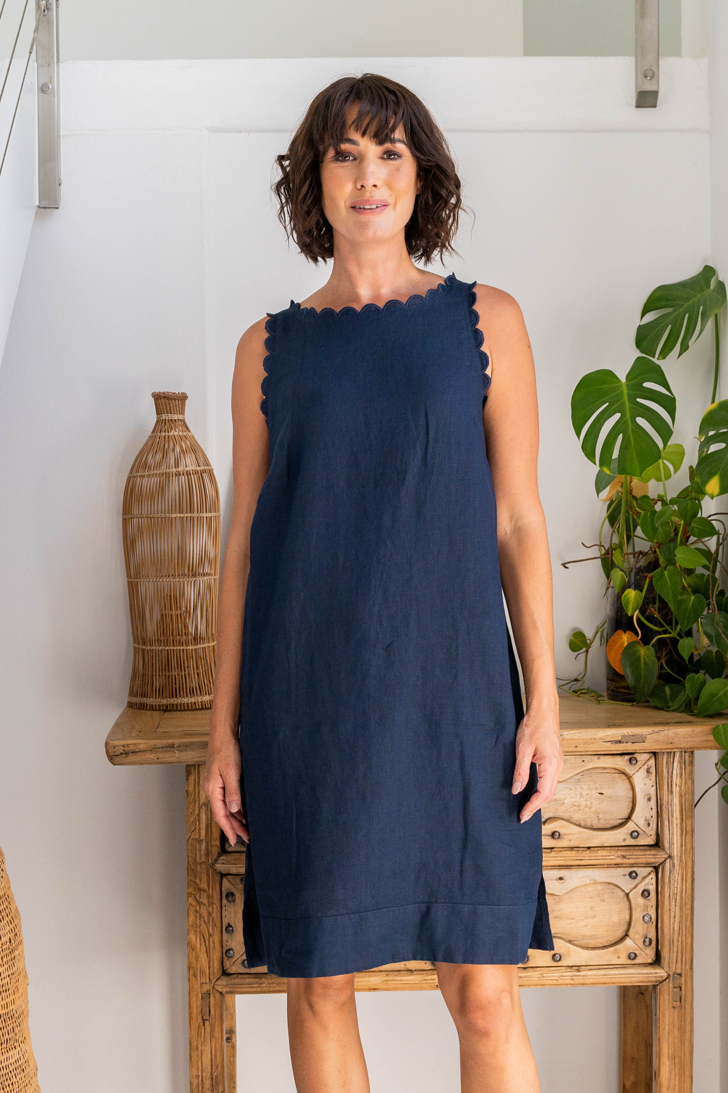 Woman wearing a navy linen See Saw dress against a white background with wooden drawers and a plant.