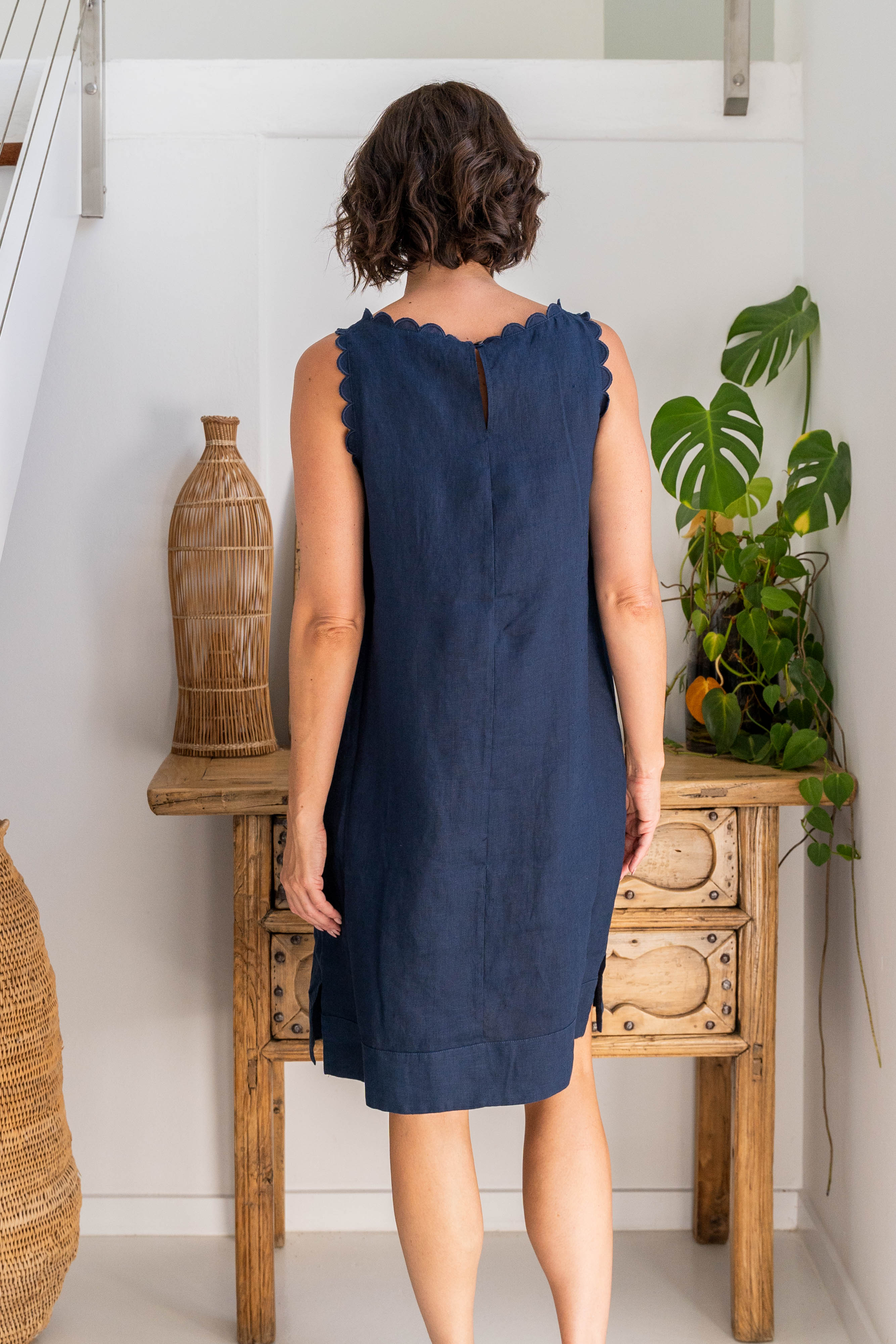 Woman wearing a navy linen See Saw dress against a white background with wooden drawers and a plant.