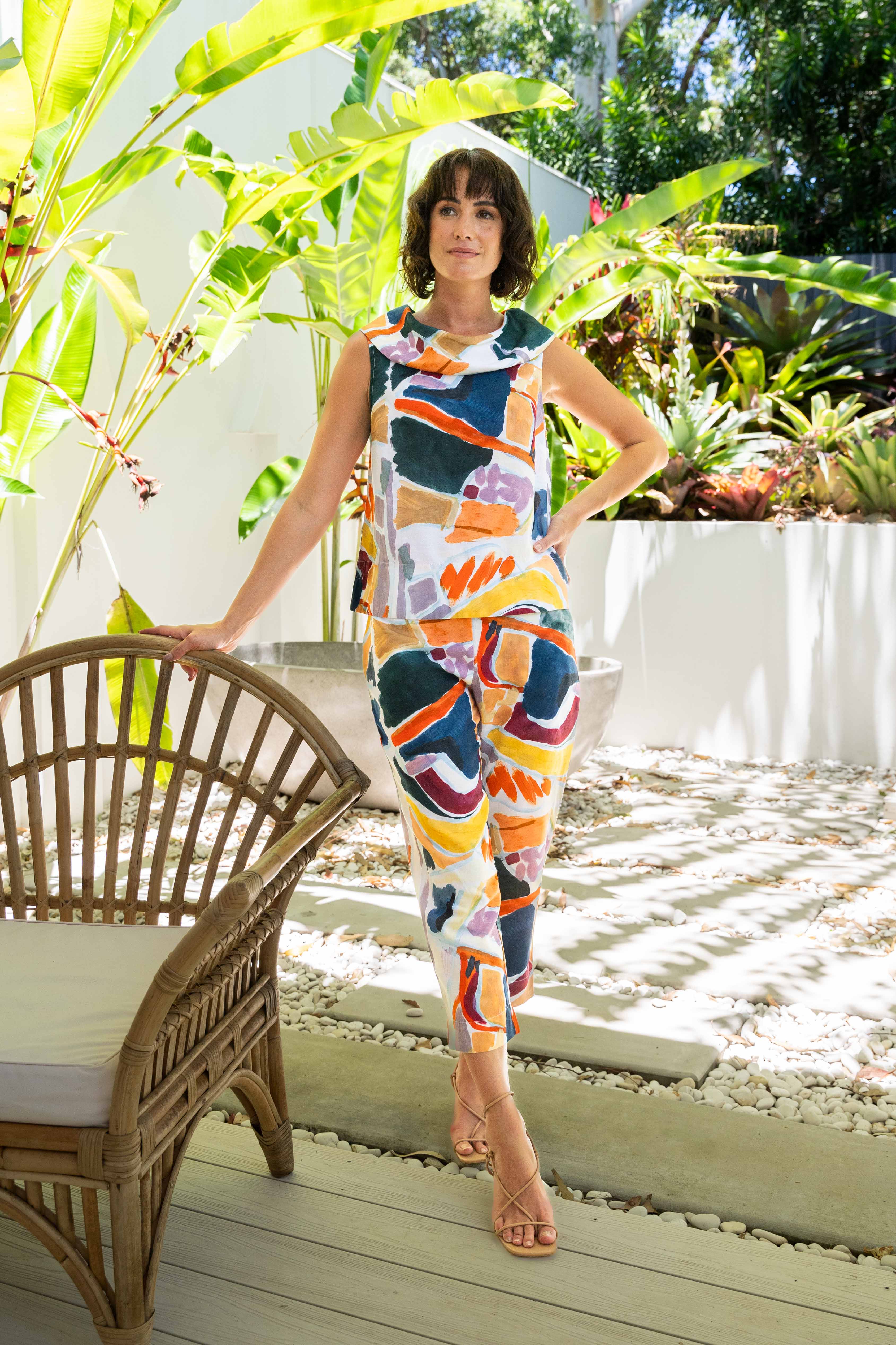 Woman in a colorful See Saw Linen pant set standing in a garden with plants and a chair.