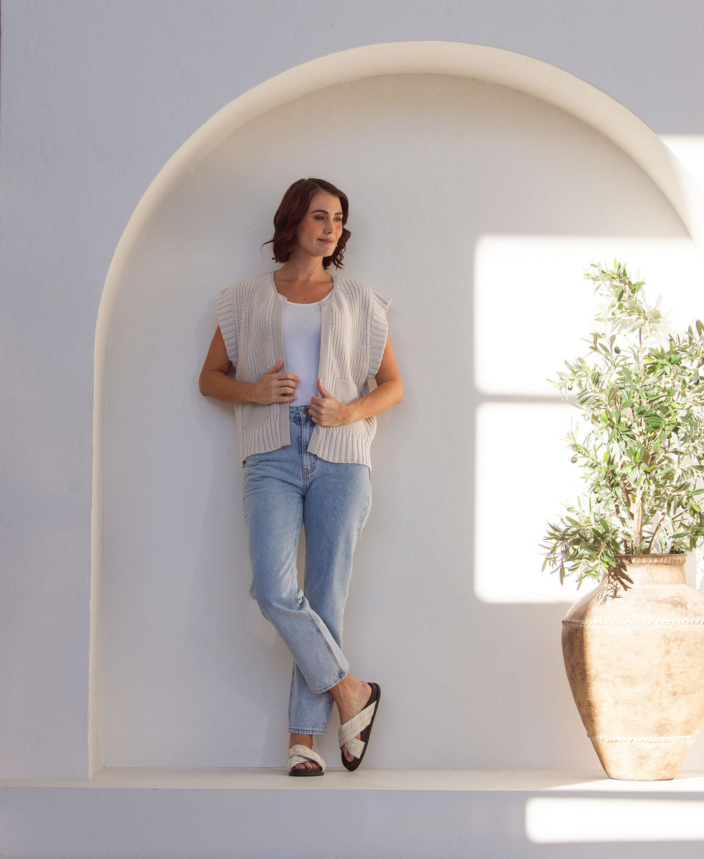 Woman wearing a stone cotton See Saw knitted vest over a white shirt and blue jeans against a white wall.