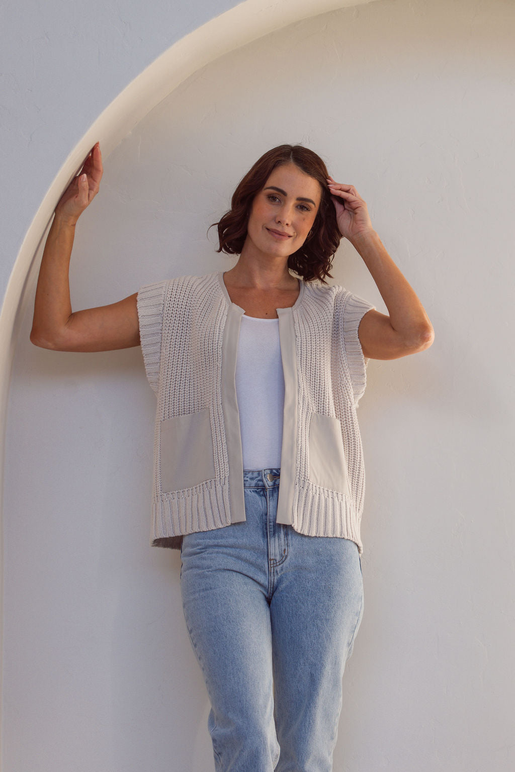 Woman wearing a stone cotton See Saw knitted vest over a white shirt and blue jeans against a white wall.