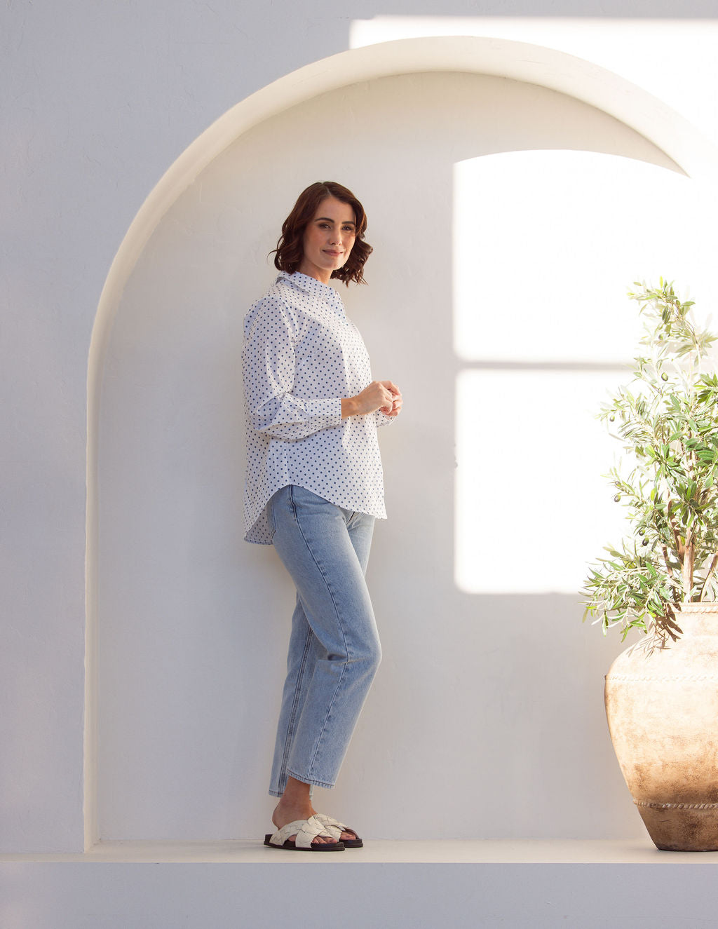 Woman wearing a classic white See Saw shirt with spot detail in a bright room with a vase and plant in the background