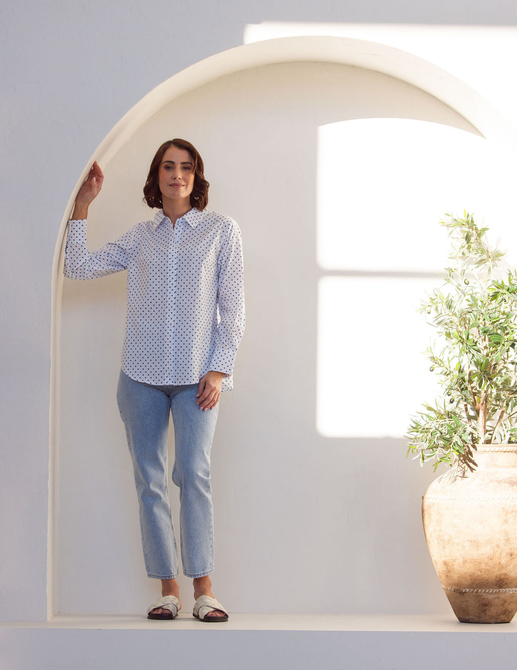 Woman wearing a classic white See Saw shirt with spot detail in a bright room with a vase and plant in the background