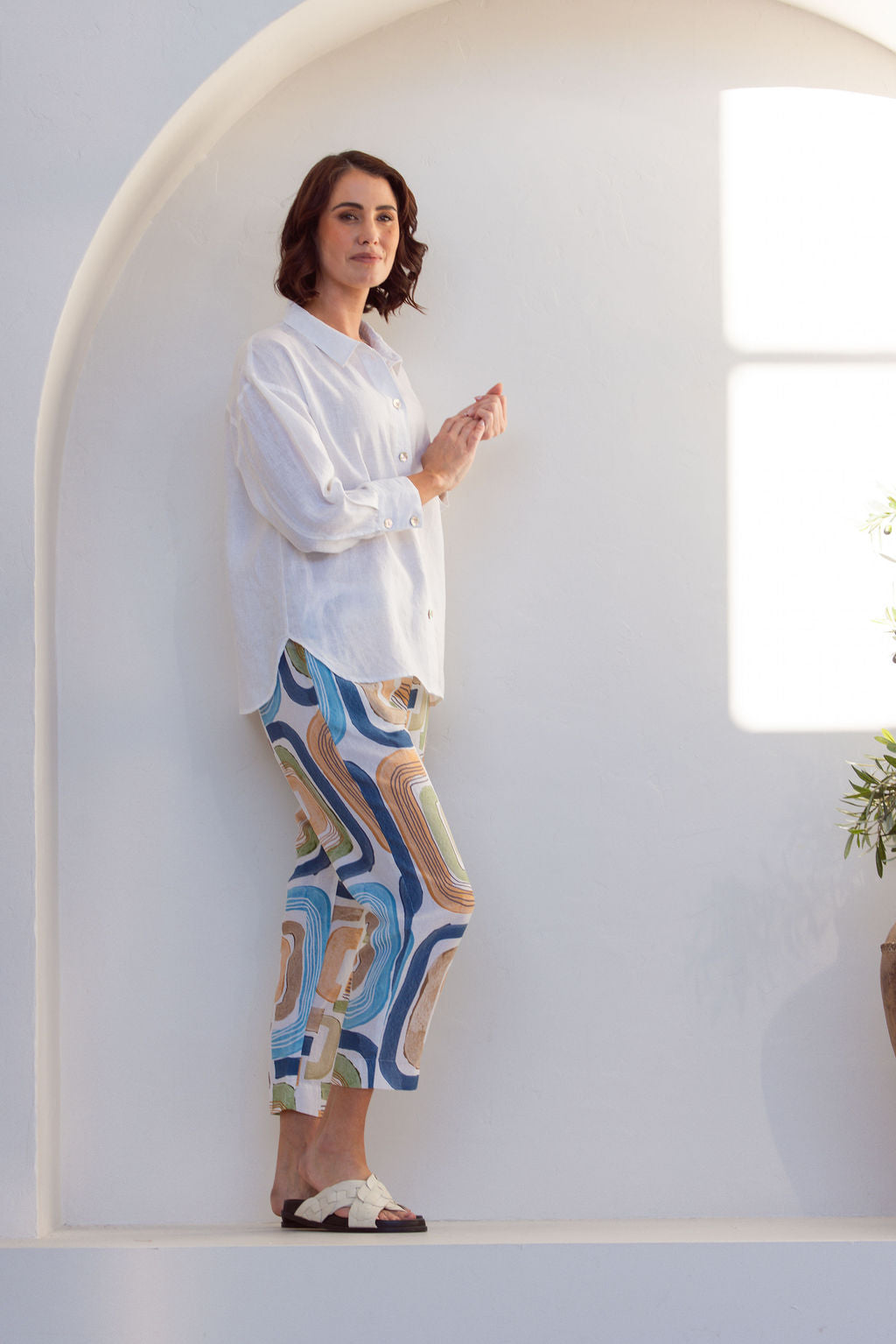 Woman wearing abstract  See Saw seascape pant and white shirt standing in a bright, sunlit room with white walls and a plant.