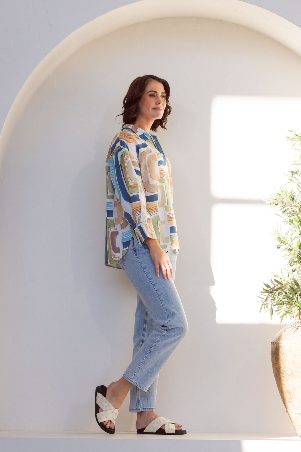 Woman wearing a long sleeve See Saw patterned linen button up shirt with jeans. Woman is standing next to a plant on a white background.