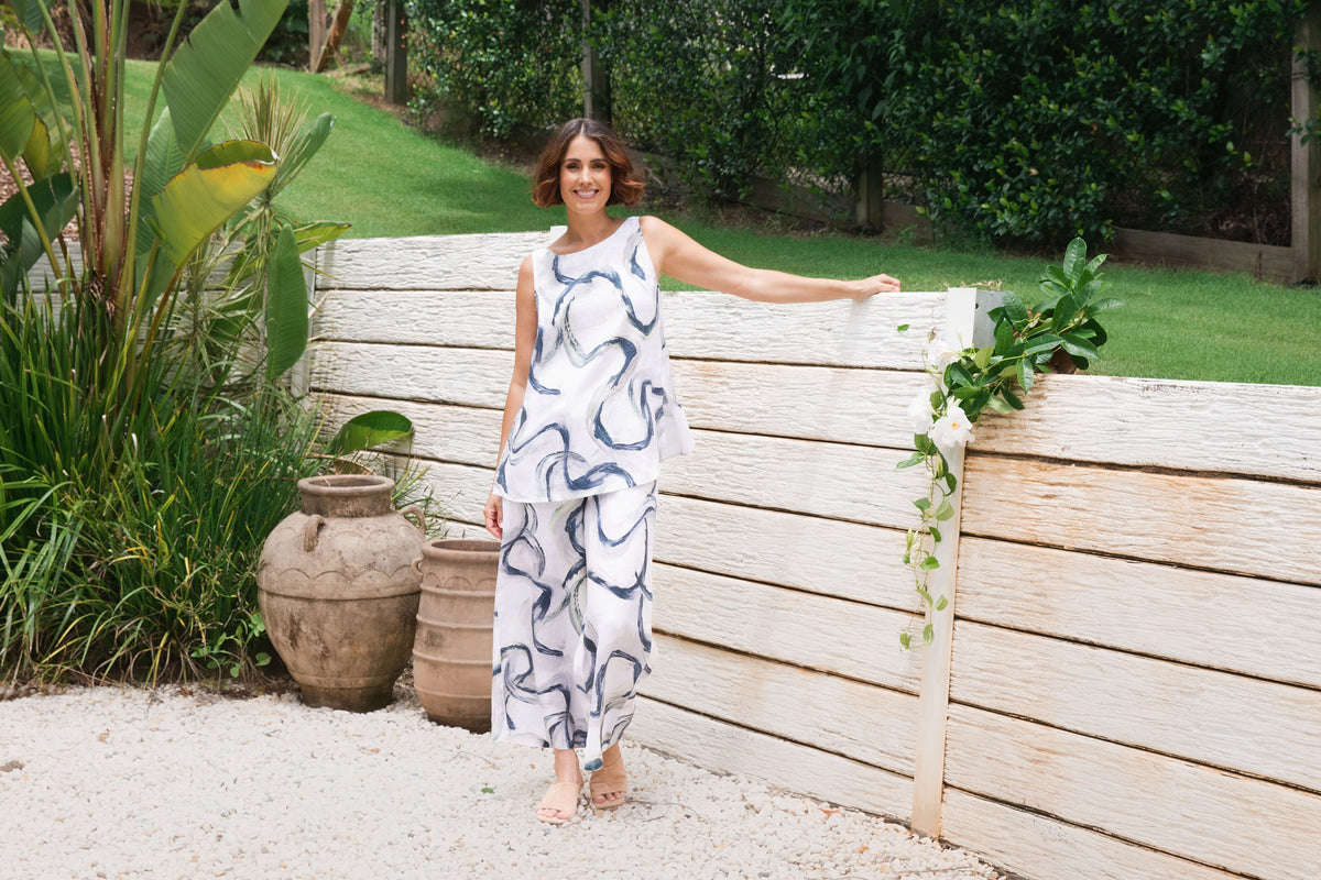 A woman stands outdoors in a garden setting next to a light-colored wooden fence, wearing a sleeveless white jumpsuit with a navy blue abstract wavy pattern. See Saw SS26 Collection