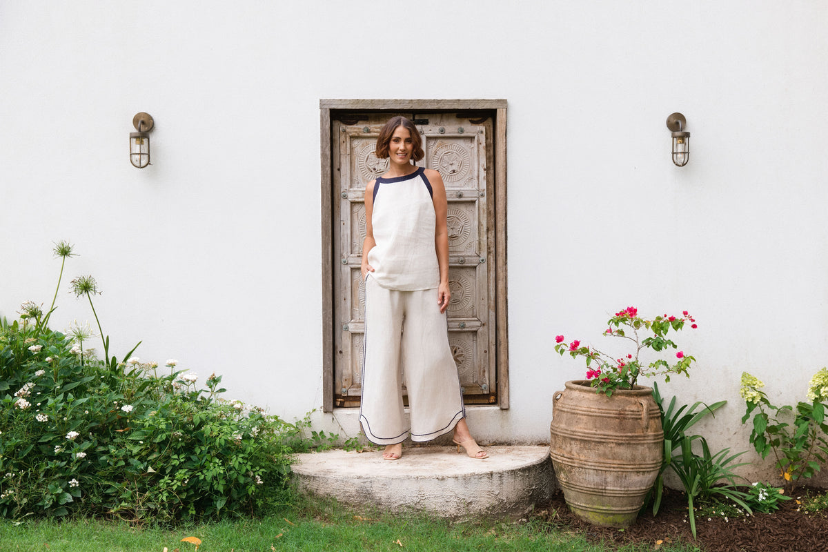 A woman stands barefoot on a stone step in front of a white stucco wall with a rustic wooden door, wearing a sleeveless cream linen top and matching wide-leg linen pants with dark blue trim. See Saw SS26 Collection