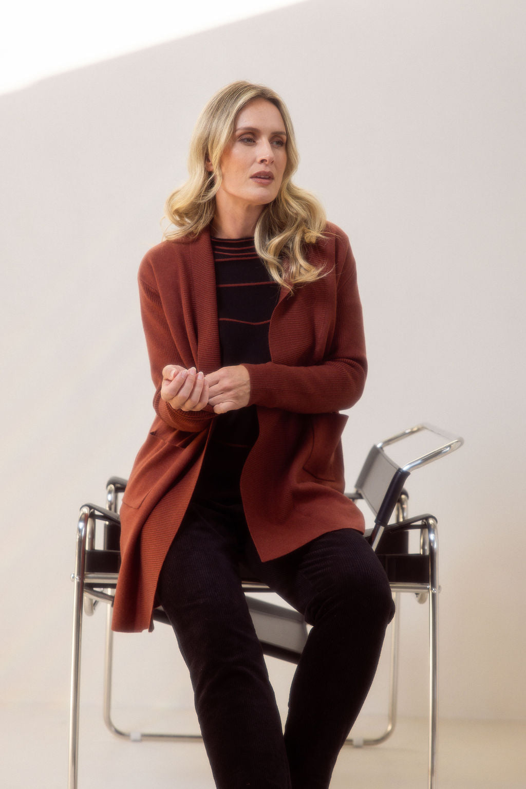 Woman wearing a brown See Saw cardigan standing next to a chair against a white background