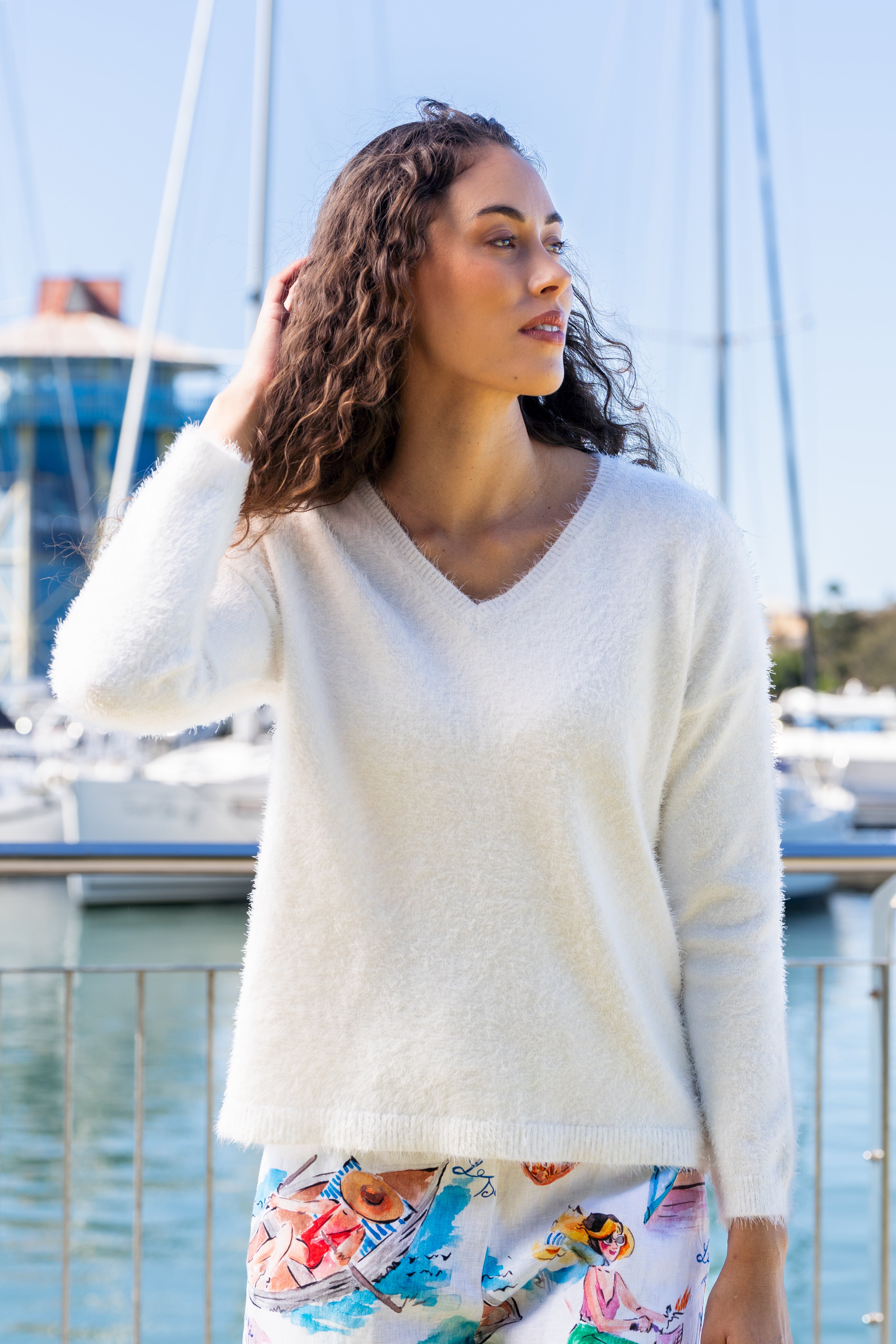 Woman wearing a white Kaleido textured sweater and colorful pants standing by a marina with boats in the background.