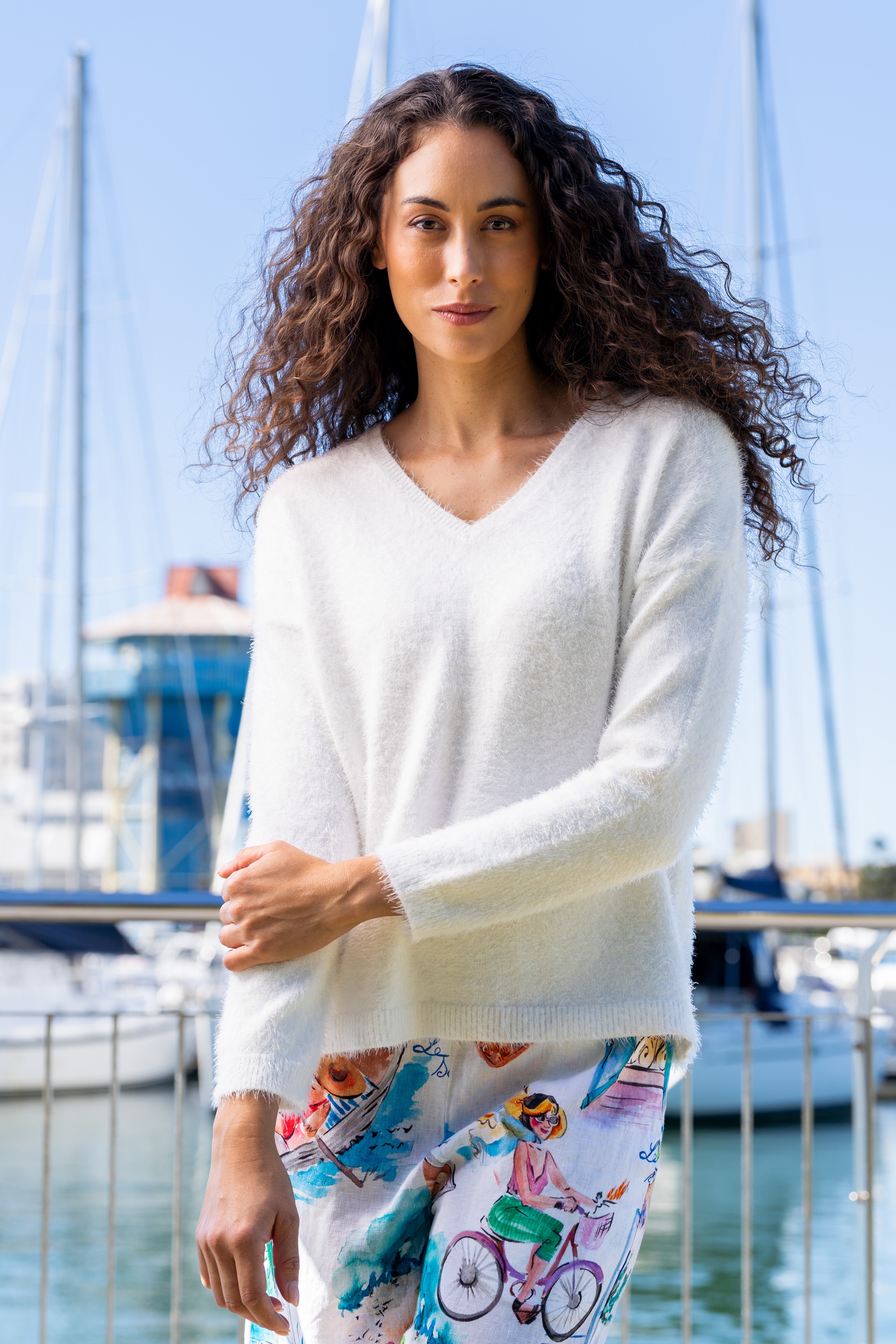 Woman wearing a white Kaleido textured sweater and colorful pants standing by a marina with boats in the background.