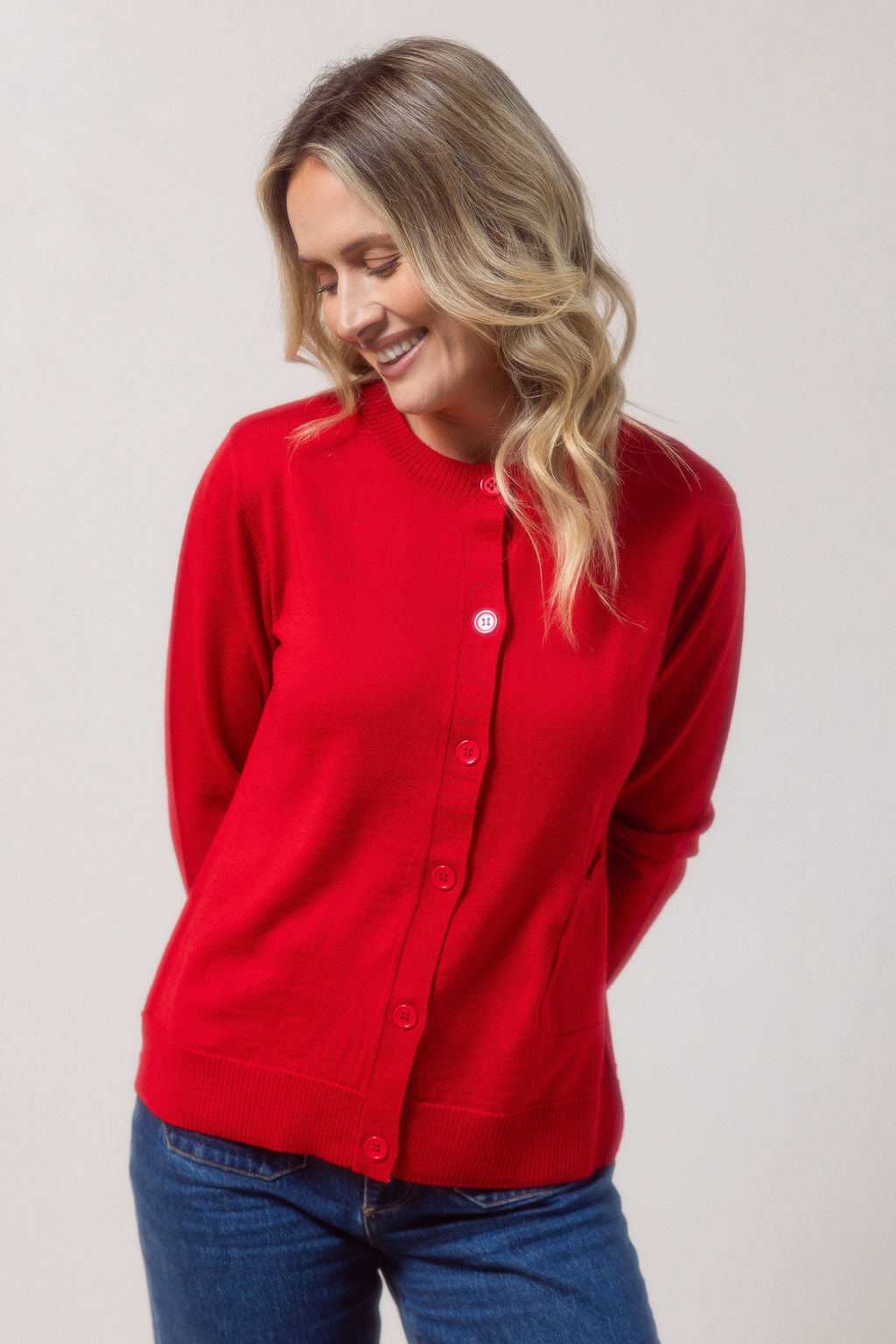 Woman wearing a merino wool See Saw red cardigan and blue jeans against a plain background