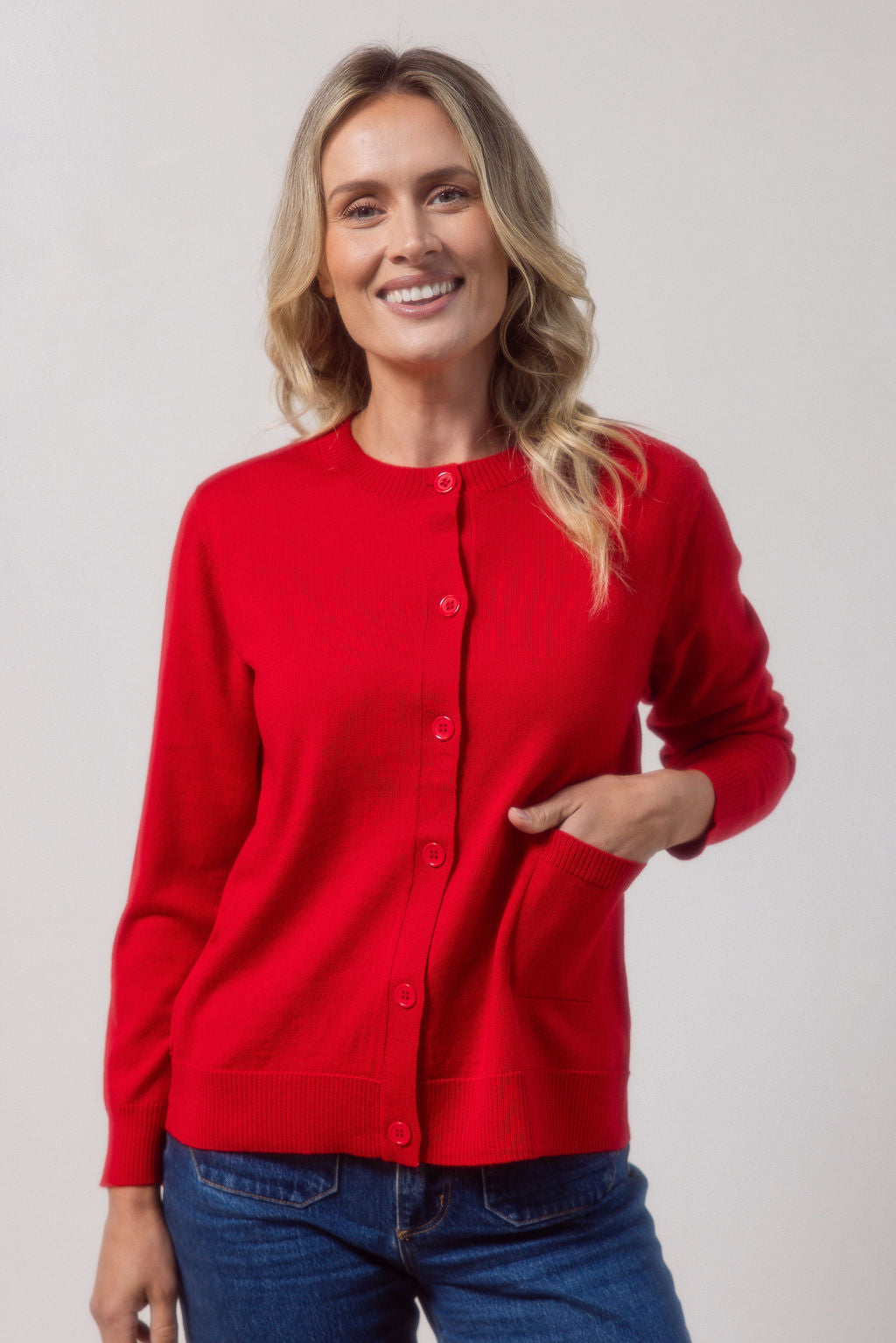 Woman wearing a merino wool See Saw red cardigan and blue jeans against a plain background