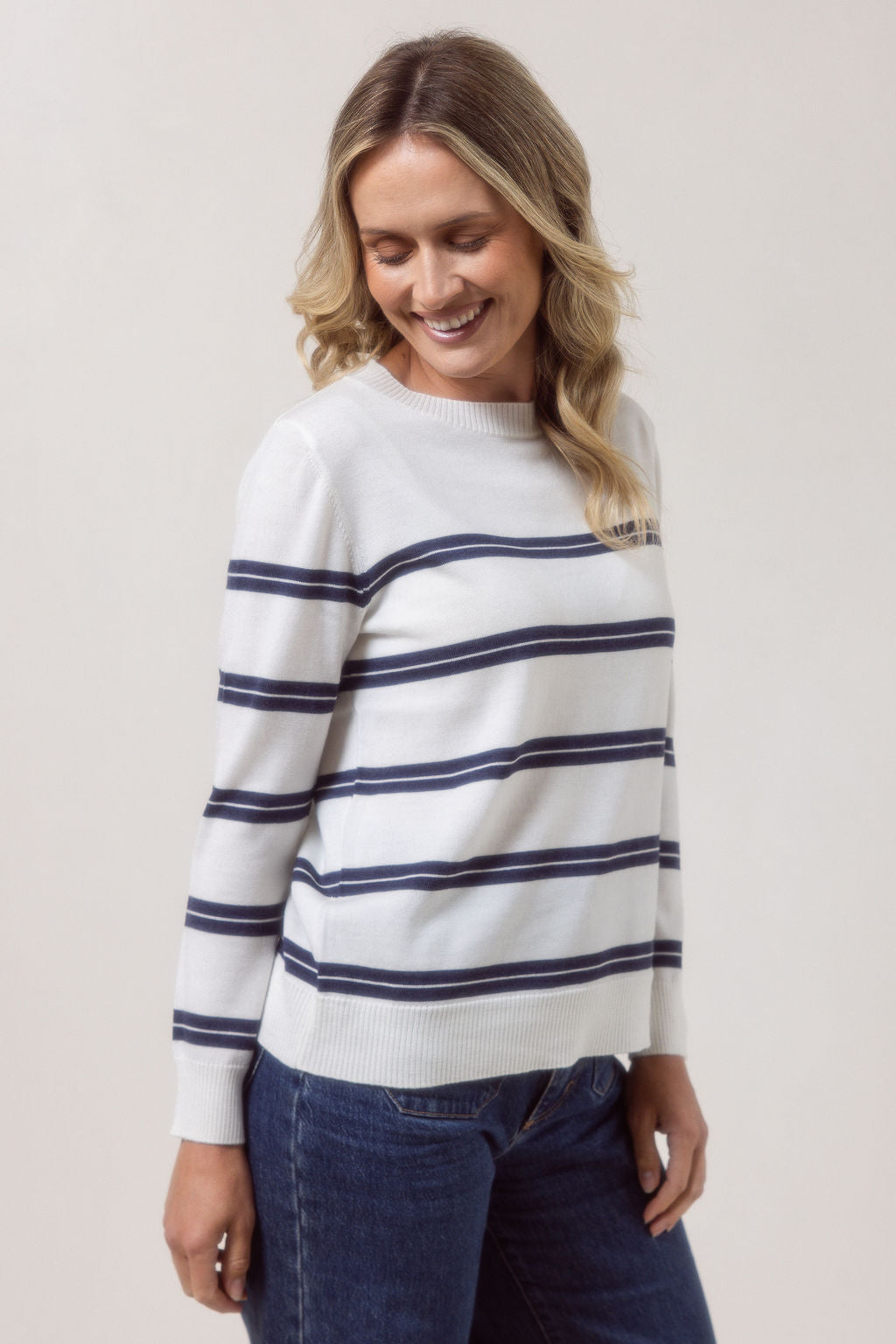 Woman wearing a white See Saw Merino wool sweater with blue stripes and dark jeans against a plain background