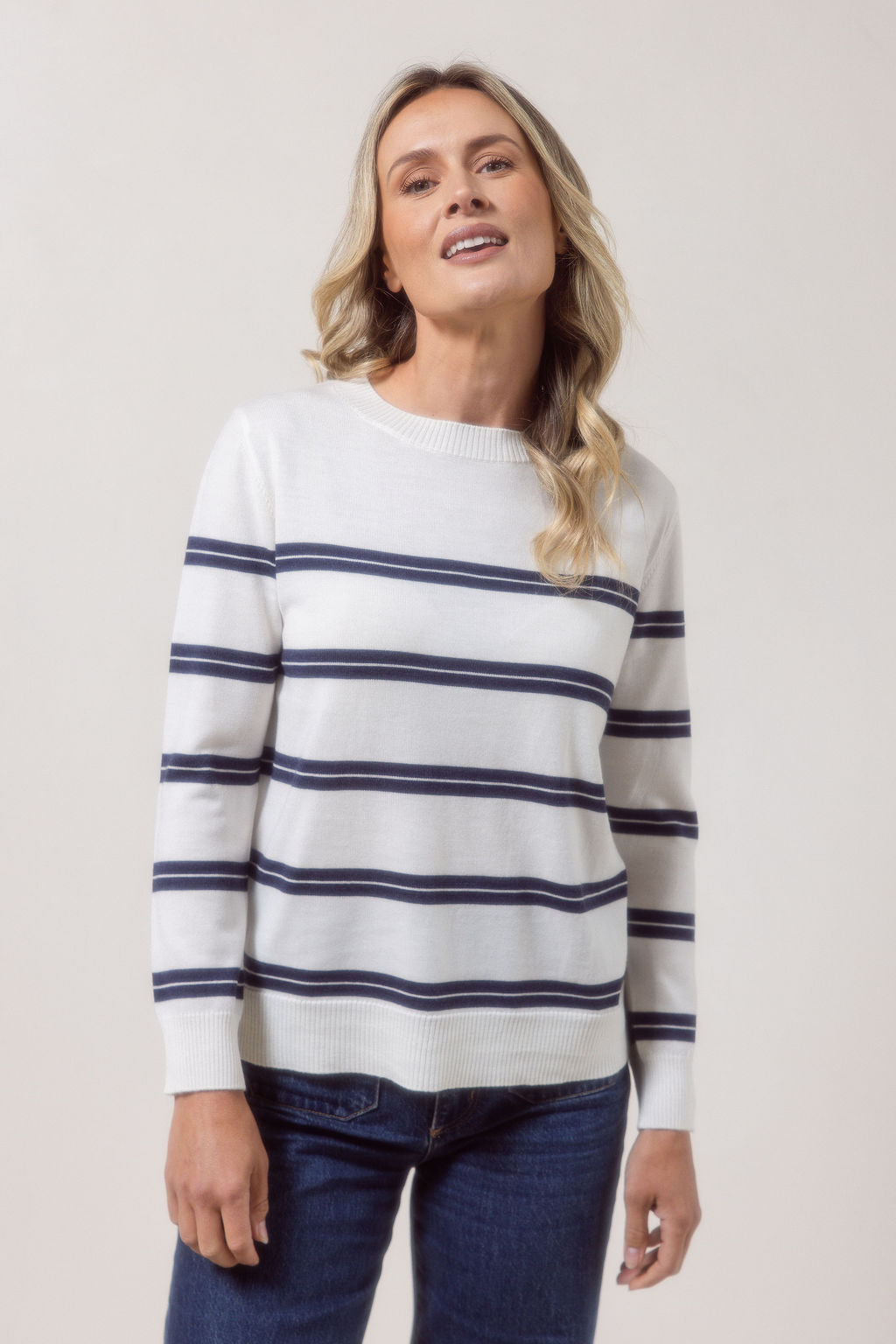 Woman wearing a white See Saw Merino wool sweater with blue stripes and dark jeans against a plain background