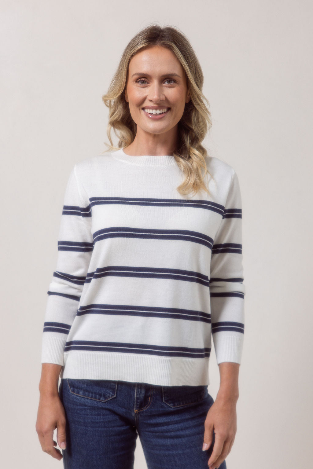 Woman wearing a white See Saw Merino wool sweater with blue stripes and dark jeans against a plain background
