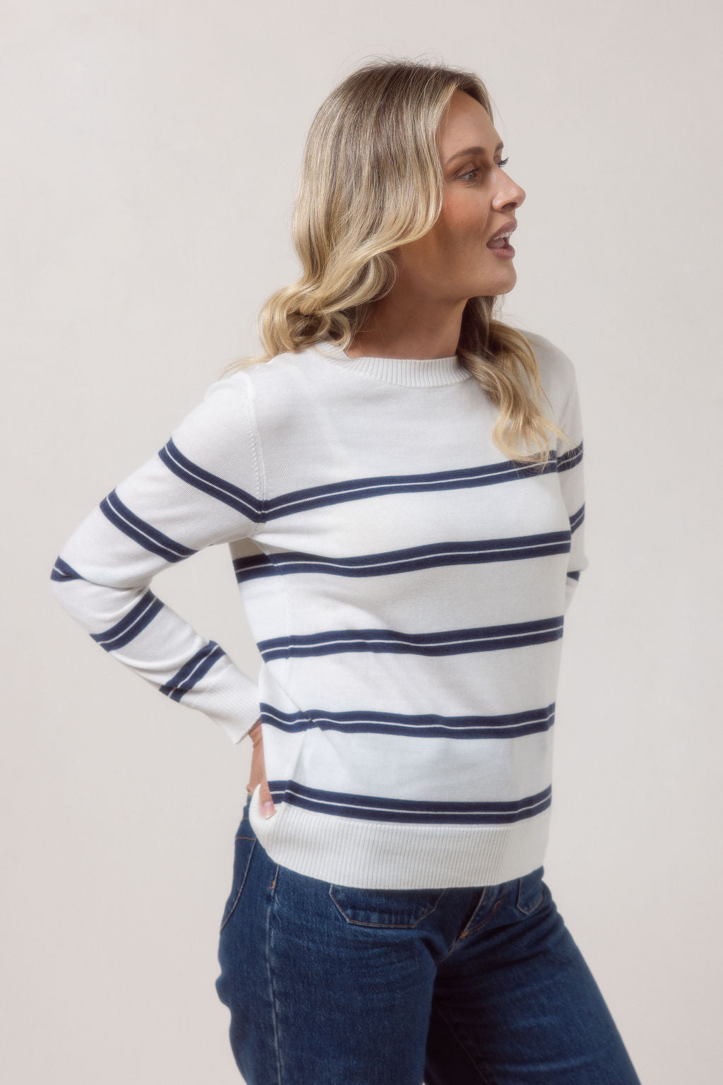 Woman wearing a white See Saw Merino wool sweater with blue stripes and dark jeans against a plain background