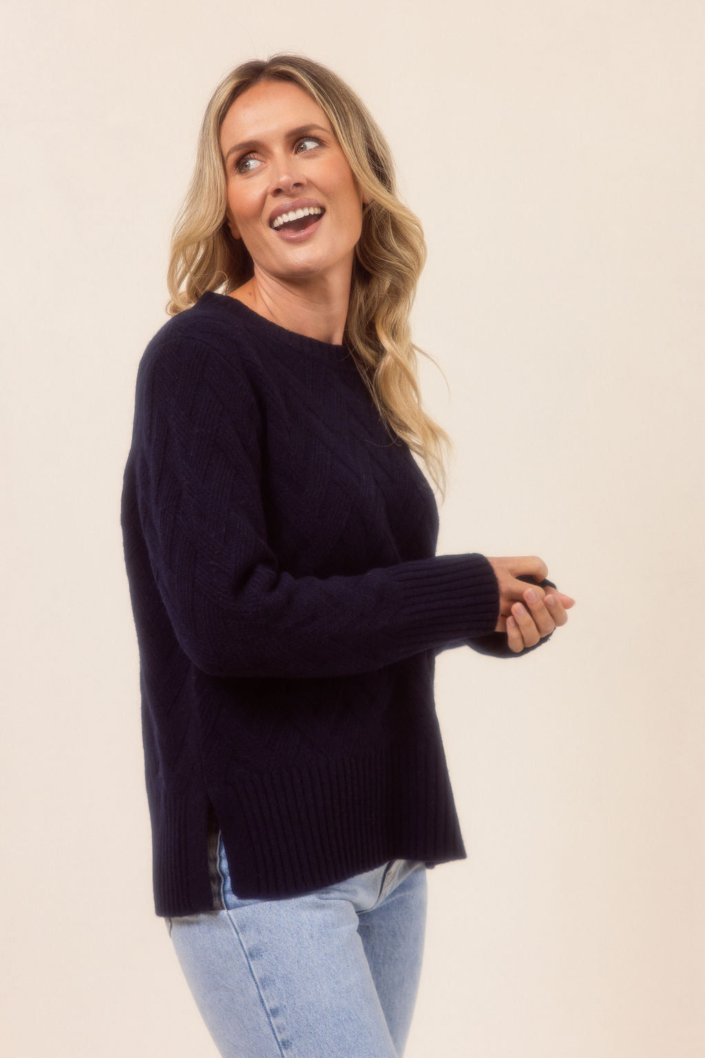 Woman wearing a navy See Saw sweater and light blue jeans against a beige background