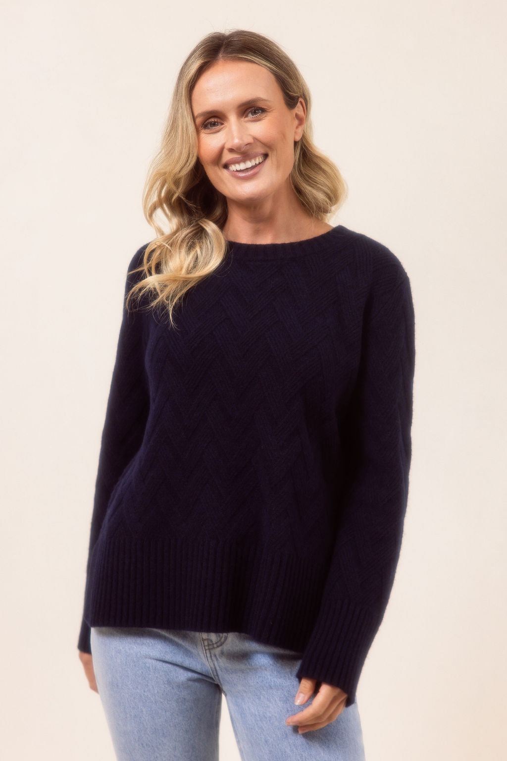 Woman wearing a navy See Saw sweater and light blue jeans against a beige background