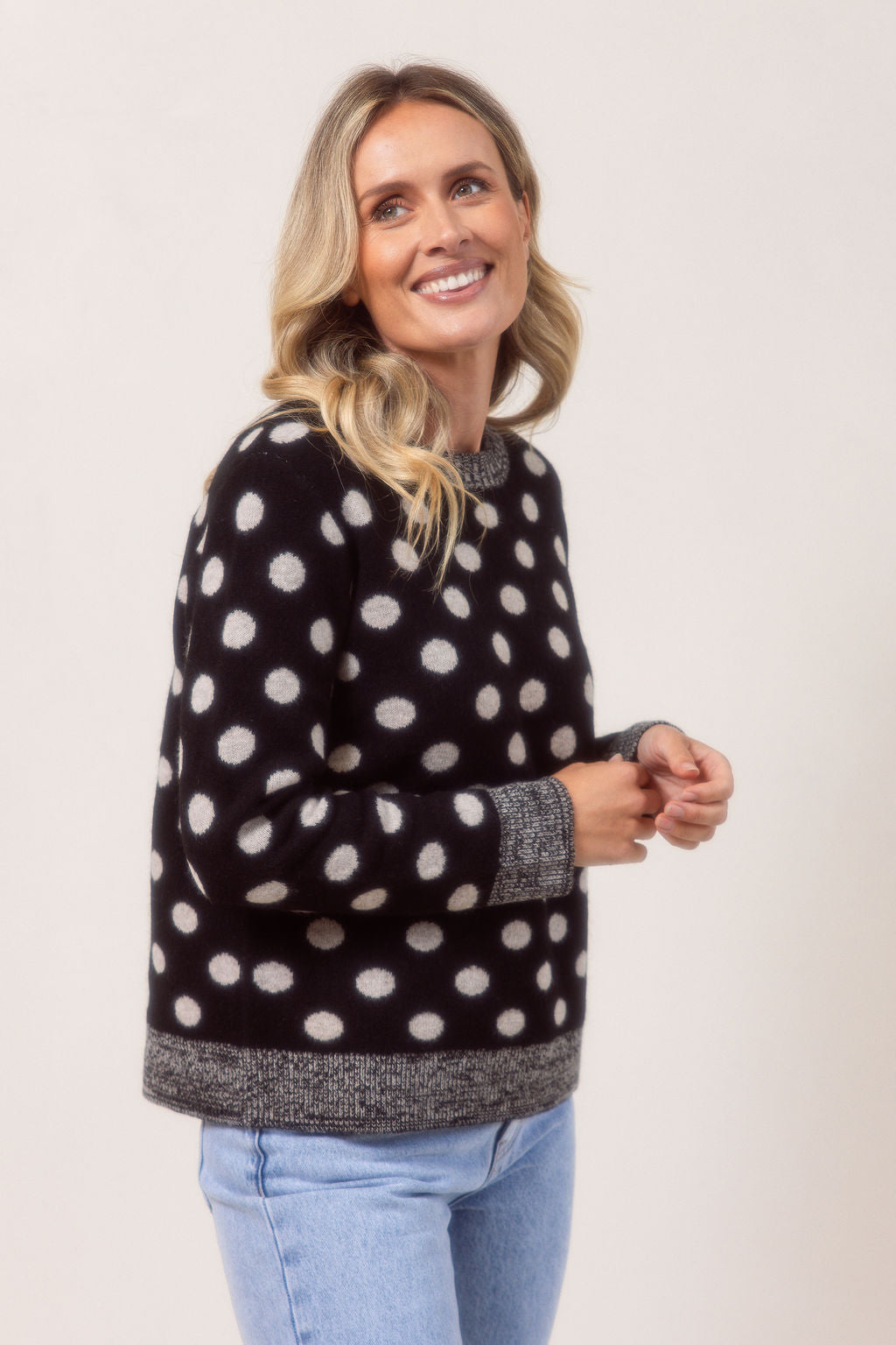 Woman wearing a See Saw Merino Wool sweater with polka dots and light blue jeans on a plain background.