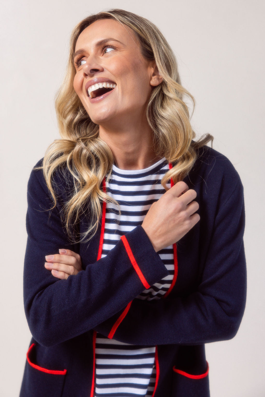 Woman wearing a See Saw navy cardigan with red trim, striped shirt, jeans, and white sneakers on a plain background