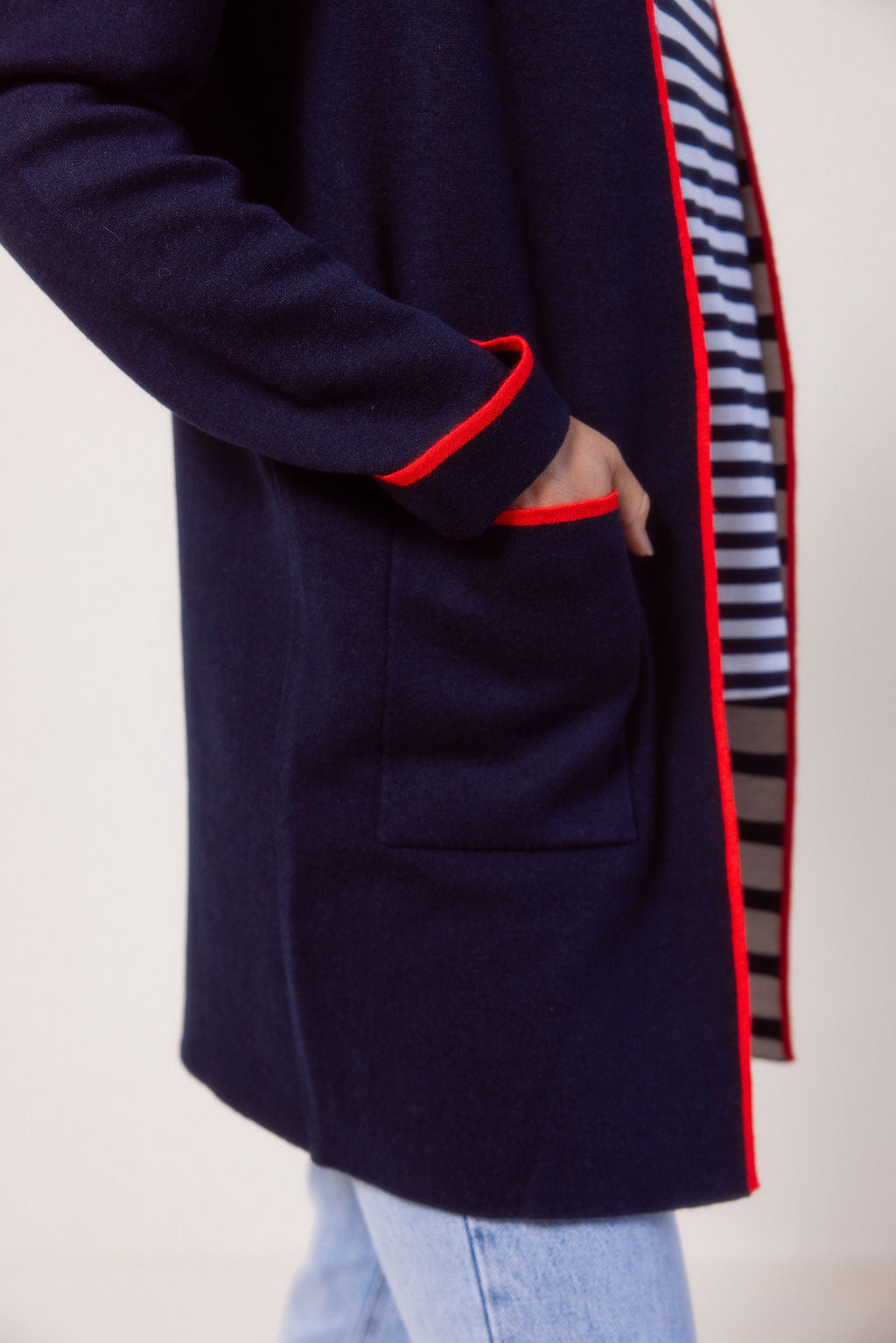 Woman wearing a See Saw navy cardigan with red trim, striped shirt, jeans, and white sneakers on a plain background